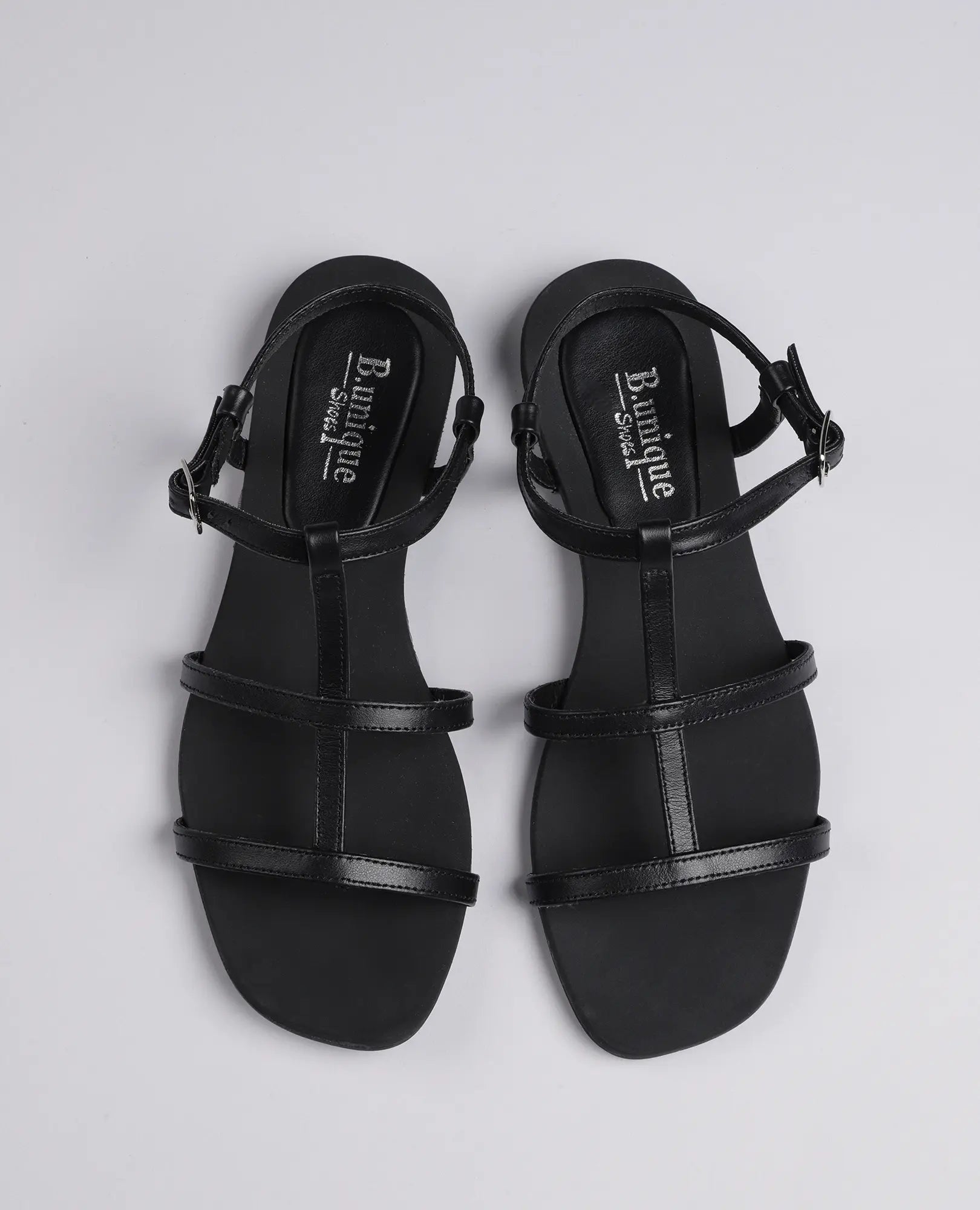 B.unique סנדלי גלדיאטור-קיילי: סנדלים שטוחים עם רצועות וסגירת אבזמים, מוצגים על רקע אפור בהיר.