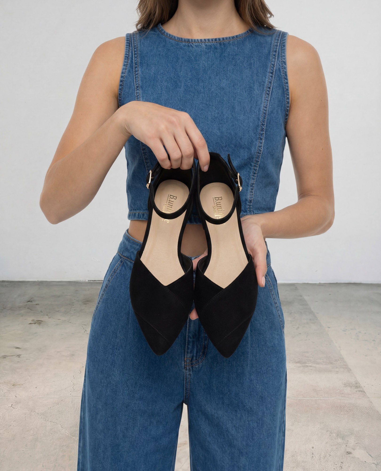 אישה לבושה בבגדי ג'ינס מחזיקה נעלי שפיץ-אלני שחורות של B.unique, בעודה עומדת בתוך הבית על רצפת בטון.