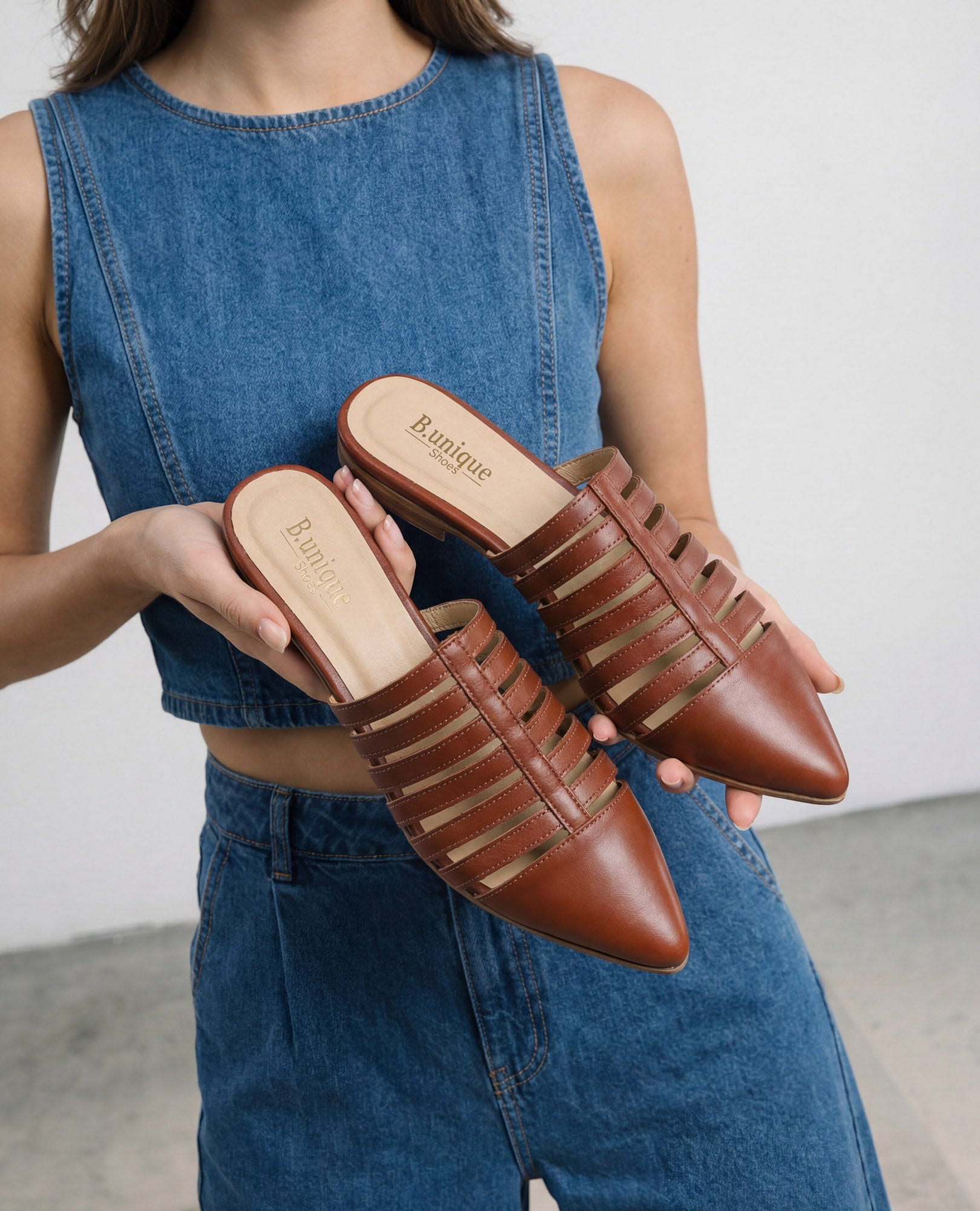 אישה לבושה בג'ינס מחזיקה כפכפים שטוחים מדגם "ליב" של המותג B.unique בצבע חום, עם שם המותג על רפידת הנעל.