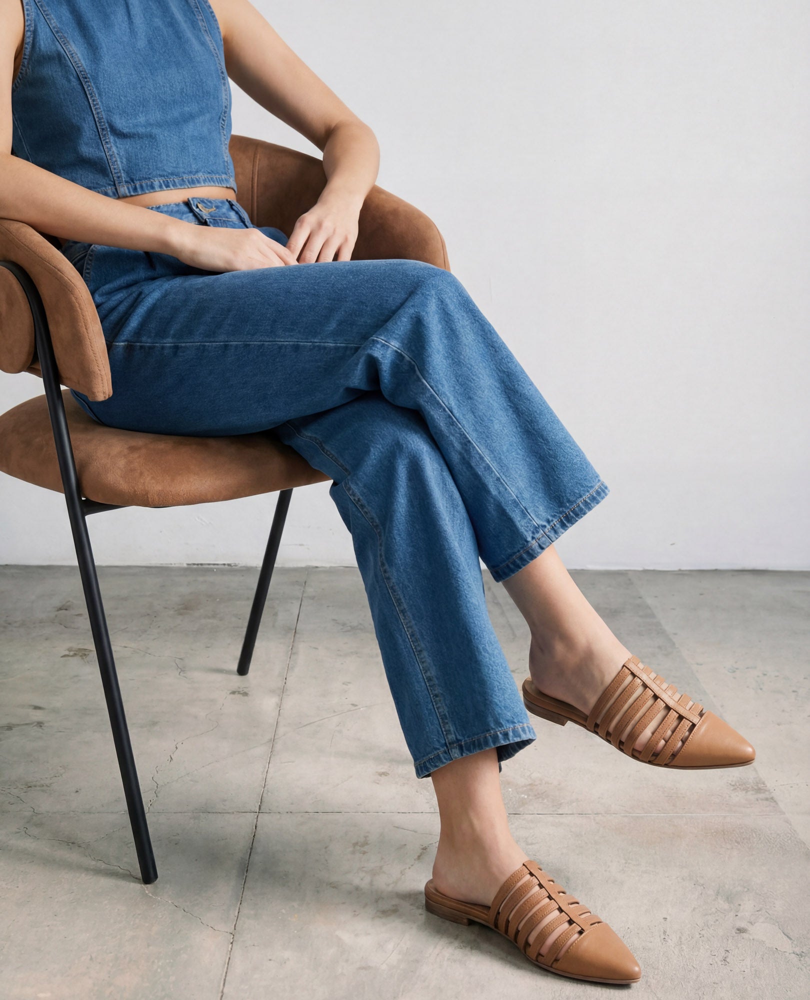 אדם לבוש בסרבל ג'ינס יושב על כיסא חום, נועל כפכפים שטוחים מדגם "ליב" של B.unique בצבע קאמל.