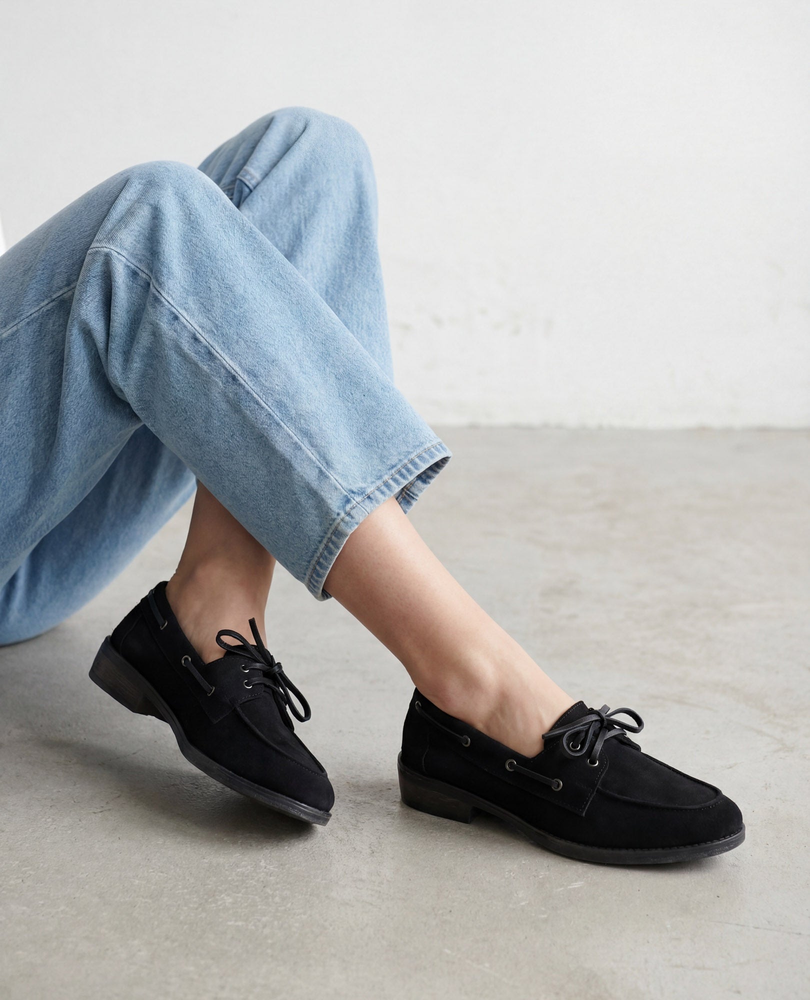 אדם לבוש בג'ינס תכלת ונעלי מוקסינים של B.unique בצבע שחור, יושב על רצפה אפורה בהירה.