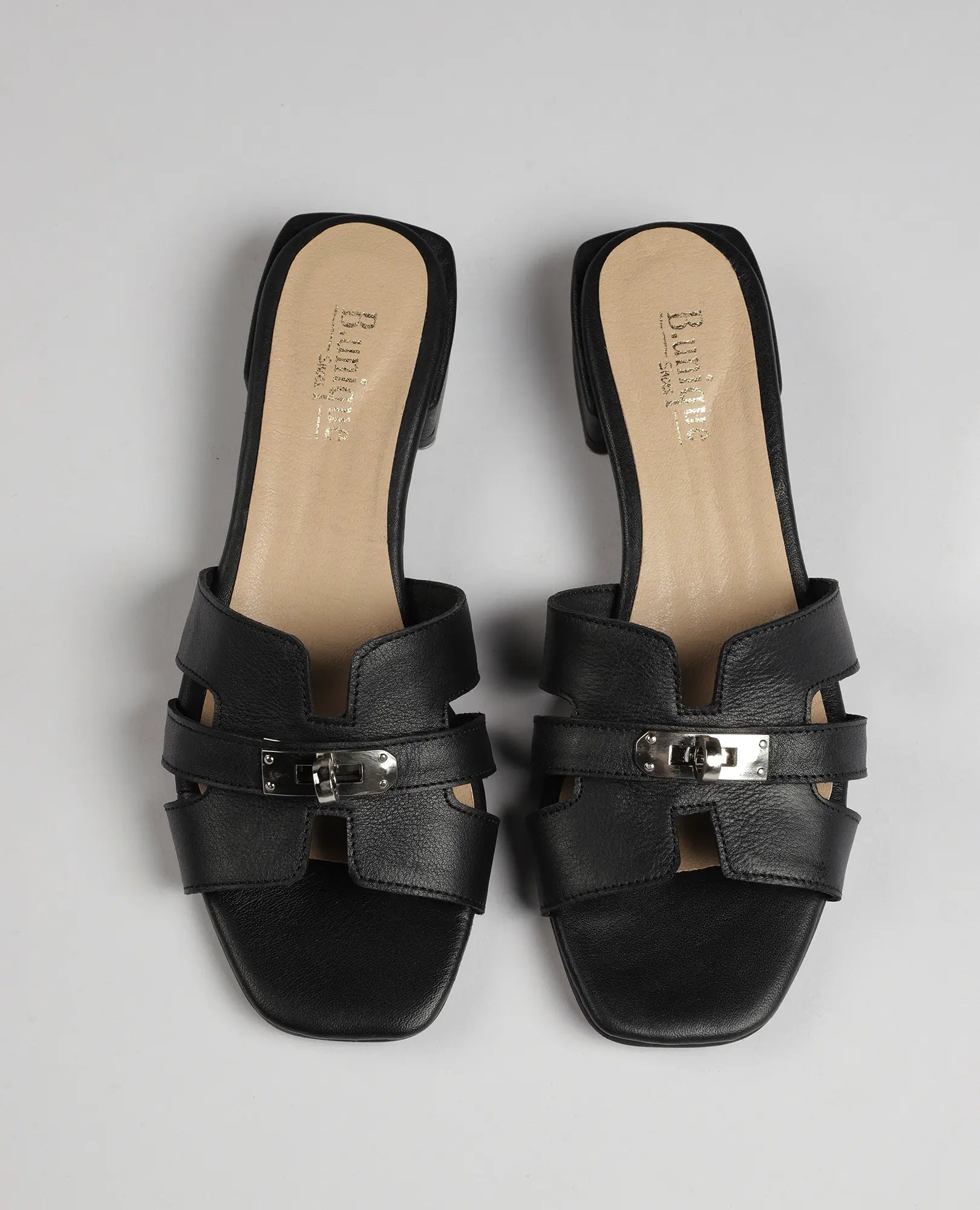 כפכפי רצועות עם אבזם – דגם "נעה" של B.unique מעור שחור עם עקב נמוך ורפידה בצבע בז', מוצגים על רקע בהיר.