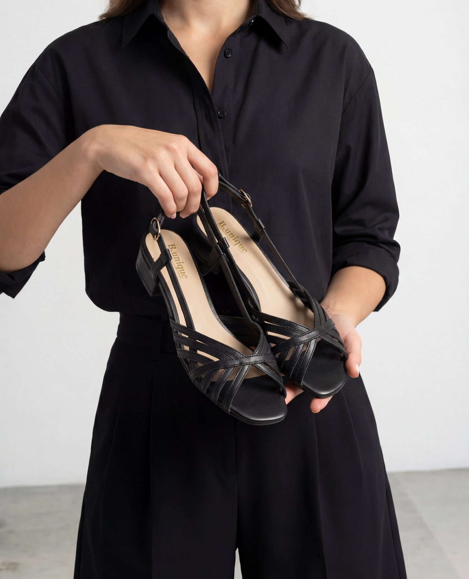 אדם מחזיק את "סנדלי עקב - שרלין" מבית B.unique בצבע שחור על רקע אחיד.