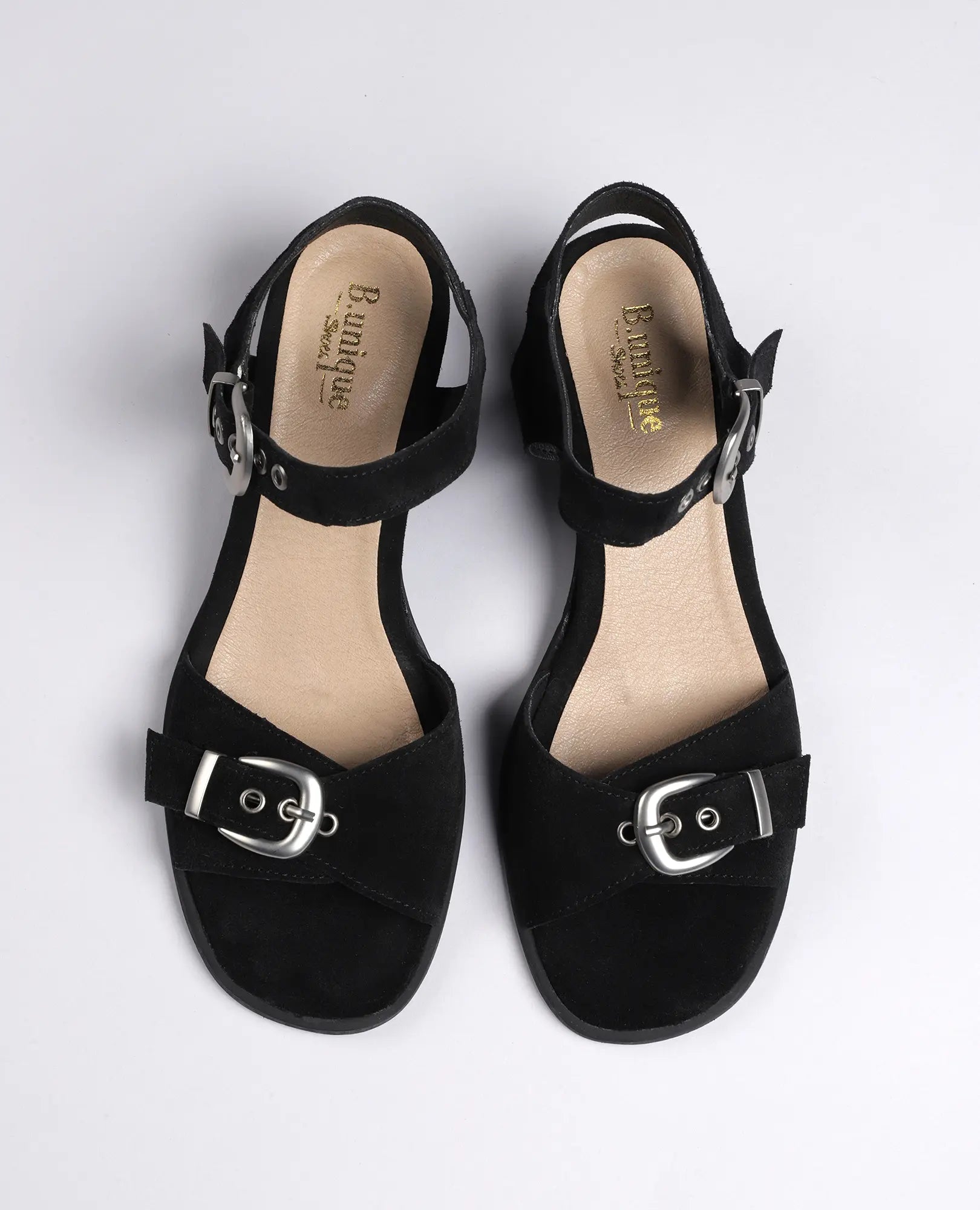 זוג סנדלים קלאסיים מדגם "קרולין" של B.unique, מעור זמש שחור, עם רצועות בקרסול ואבזמים כסופים על רקע בהיר.