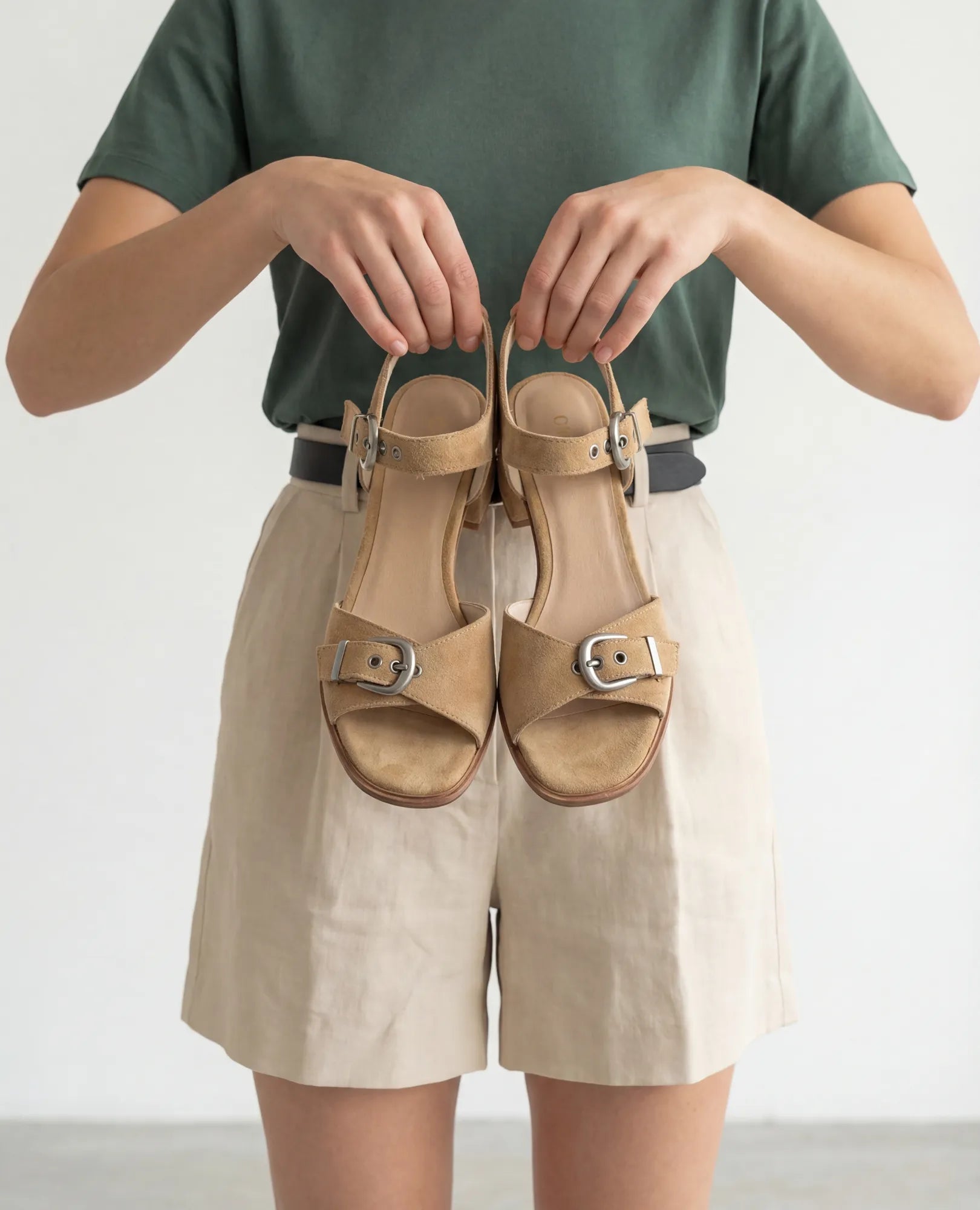 אדם לבוש בחולצה ירוקה ובמכנסיים קצרים בצבע בז' מחזיק בשתי ידיו את "סנדלים קלאסיים - קרולין" מבית B.unique.