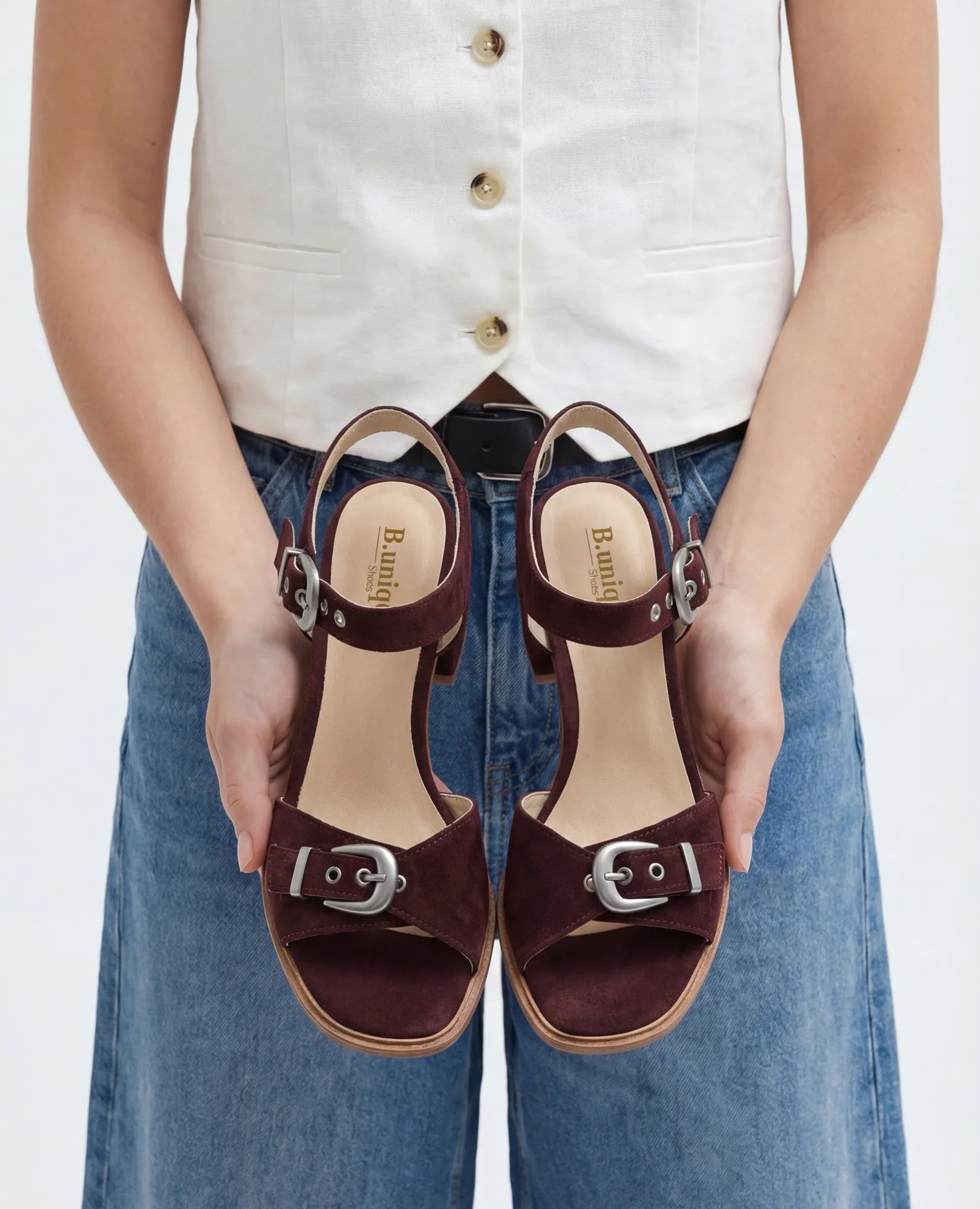 אדם לבוש בג'ינס ובגופייה לבנה מחזיק את הסנדלים הקלאסיים של B.unique – קרולין, ופונה למצלמה.