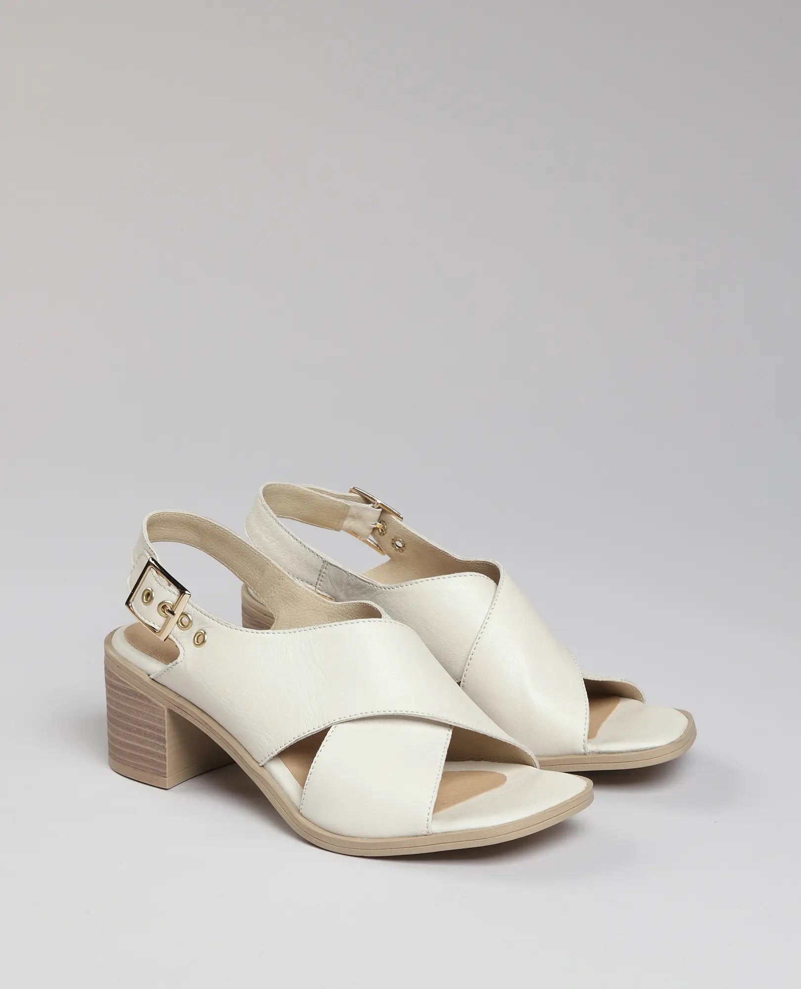 זוג סנדלי עקב לבנים מדגם "עיינה" מבית B.unique, עם רצועות מצטלבות ורצועת קרסול עם אבזם על רקע בהיר.