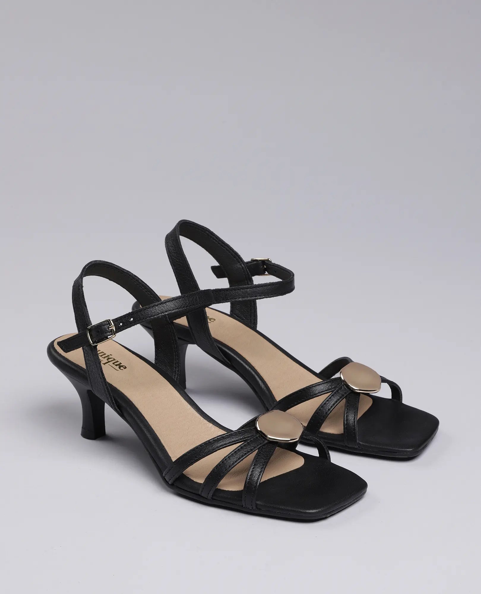 נעלי עקב דליה מבית B.unique בעיצוב שחור אלגנטי עם רצועות דקות ועיטורי זהב אובליים בחלק הקדמי.