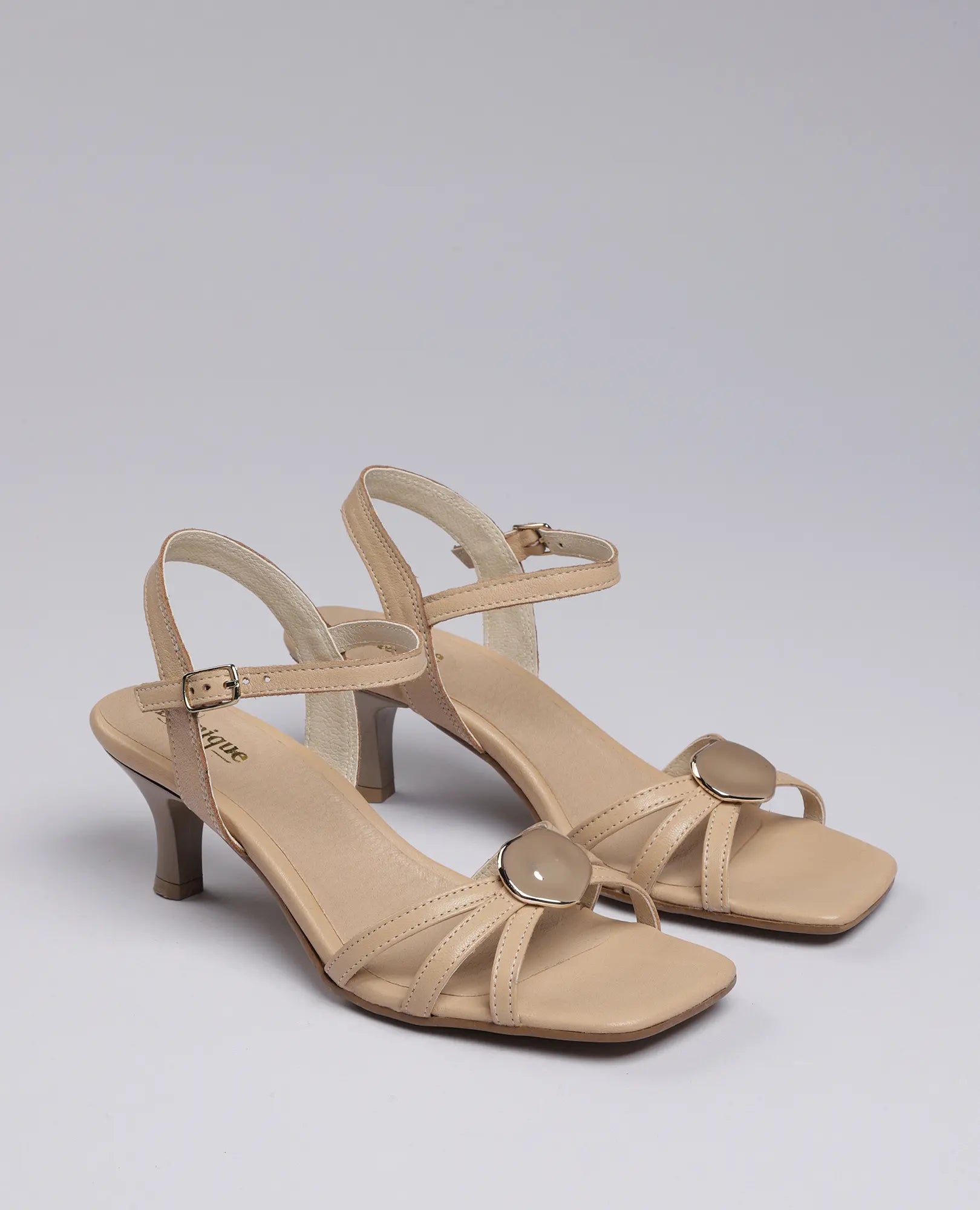 סנדל עקב - דגם "דיליה" של B.unique בעיצוב בז', עם רצועת קרסול ואבזמים עגולים בחזית, מוצג על רקע חלק.