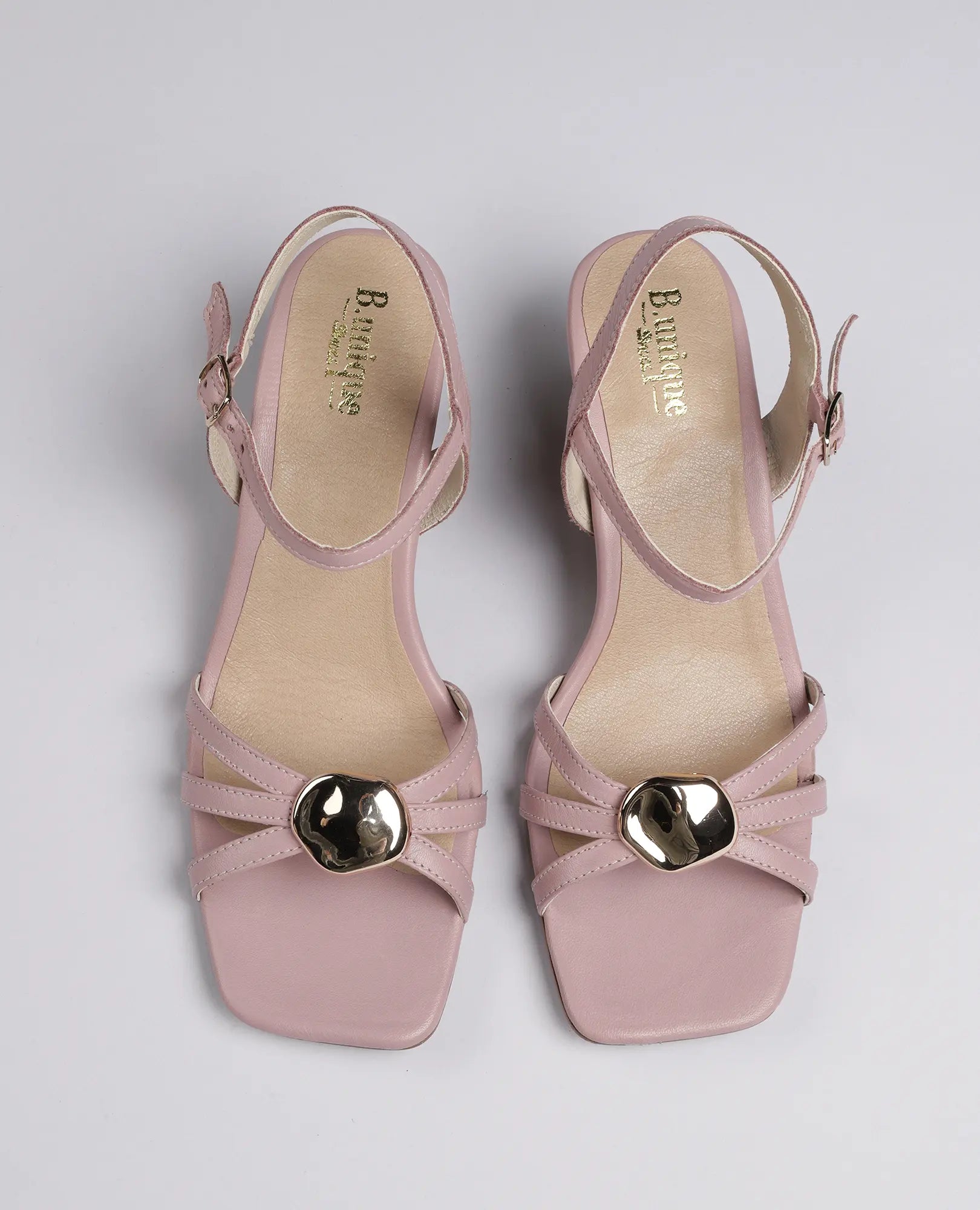 סנדל העקב "דיליה" מבית B.unique: סנדלי עור בצבע ורוד בהיר עם אבזמים מוזהבים ורצועות בקרסול, בתצוגה מלמעלה.