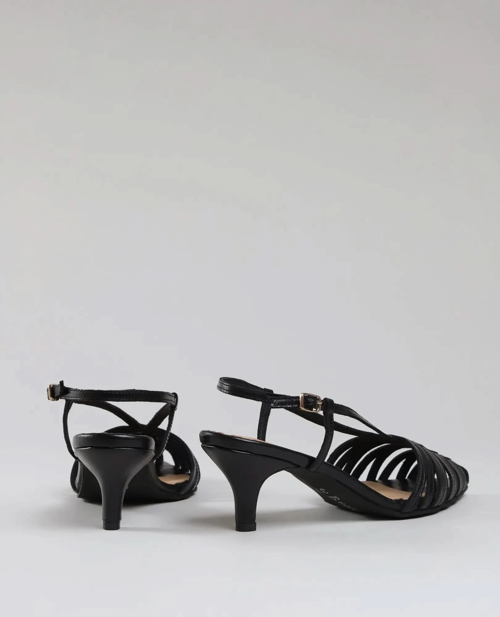 A pair of elegant black סנדלי עקב- מיכאלה with ankle straps and small buckles are displayed against a plain light gray background, shown from back and side angles.