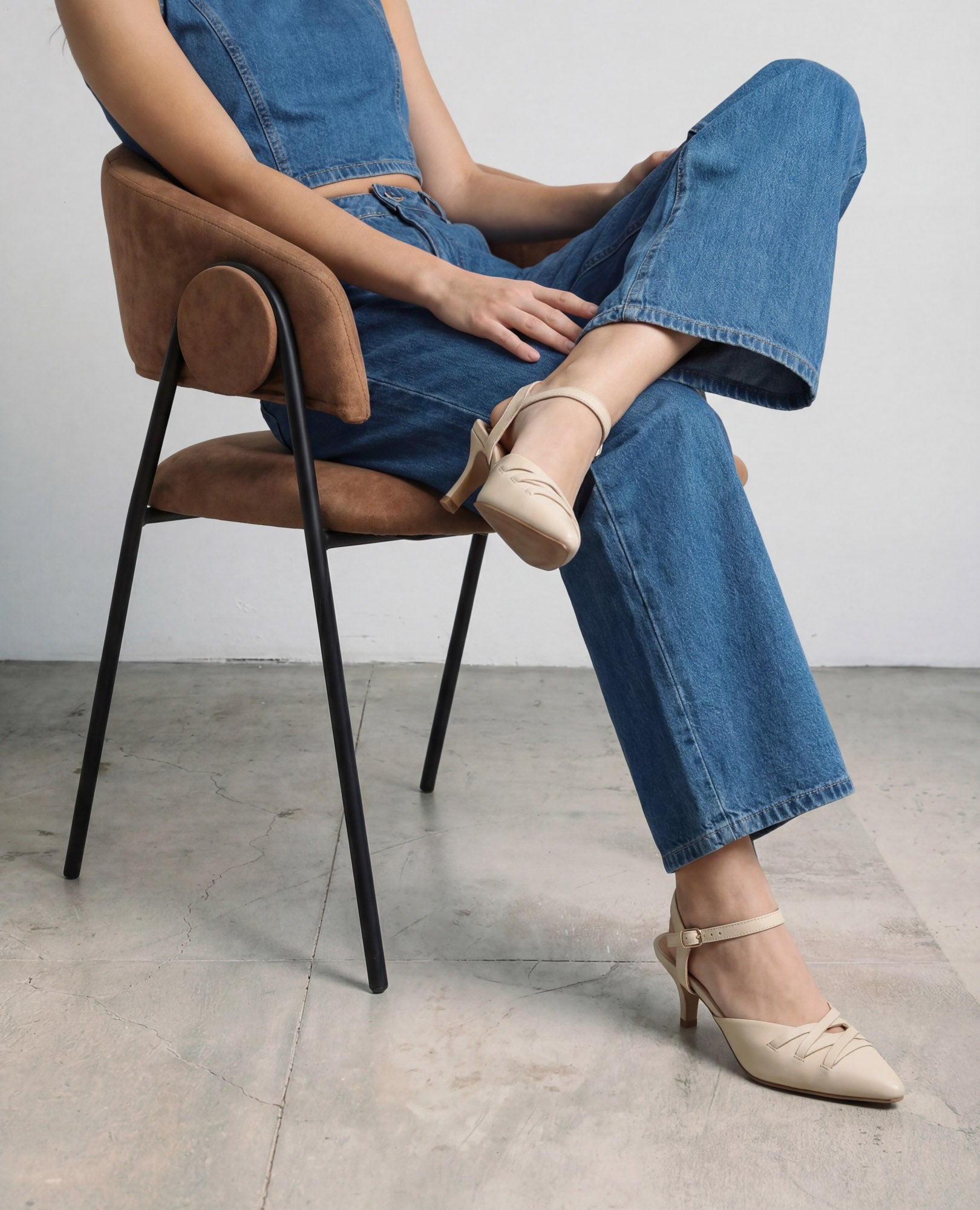 אישה לבושה בג'ינס כחול יושבת על כיסא חום, רגליה משוכלות, נועלת נעלי עקב של B.unique בצבע בז' על רצפת בטון מלוטשת.