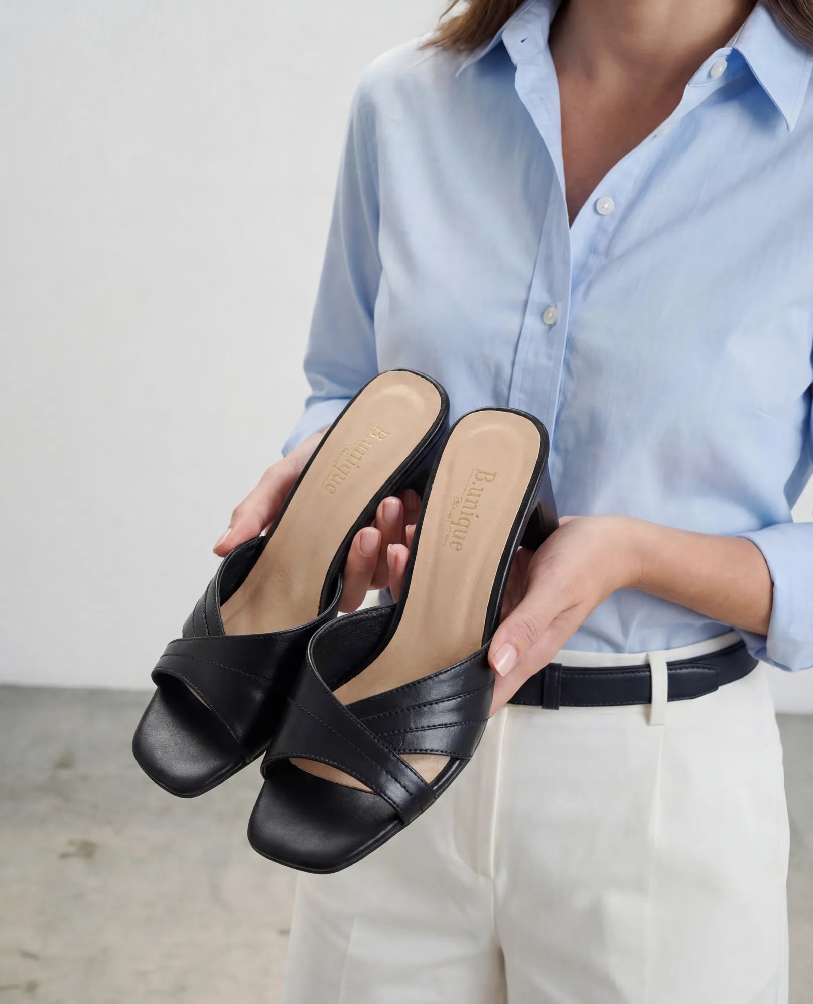 אישה לבושה בחולצה כחולה ומכנסיים לבנים מחזיקה כפכפי עקב של המותג B.unique בצבע ארגמן שחור, עם רצועות עור איכותיות בצורת שתי וערב.