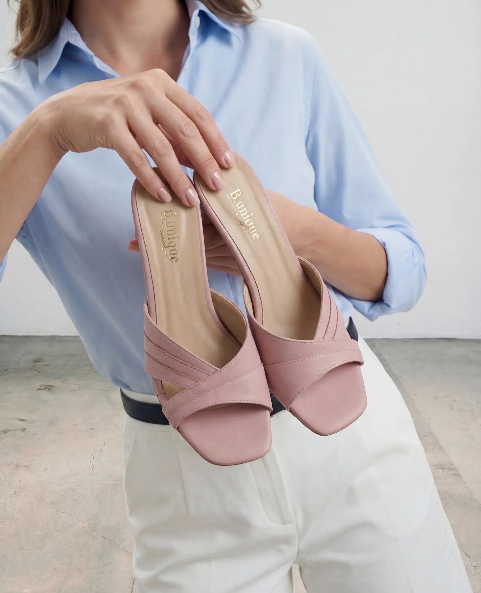 אישה לבושה בחולצה כחולה ומכנסיים לבנים אוחזת בסנדלי עקב של המותג B.unique – סנדלים בצבע ארגמן ורוד בהיר עם רצועות מצטלבות.