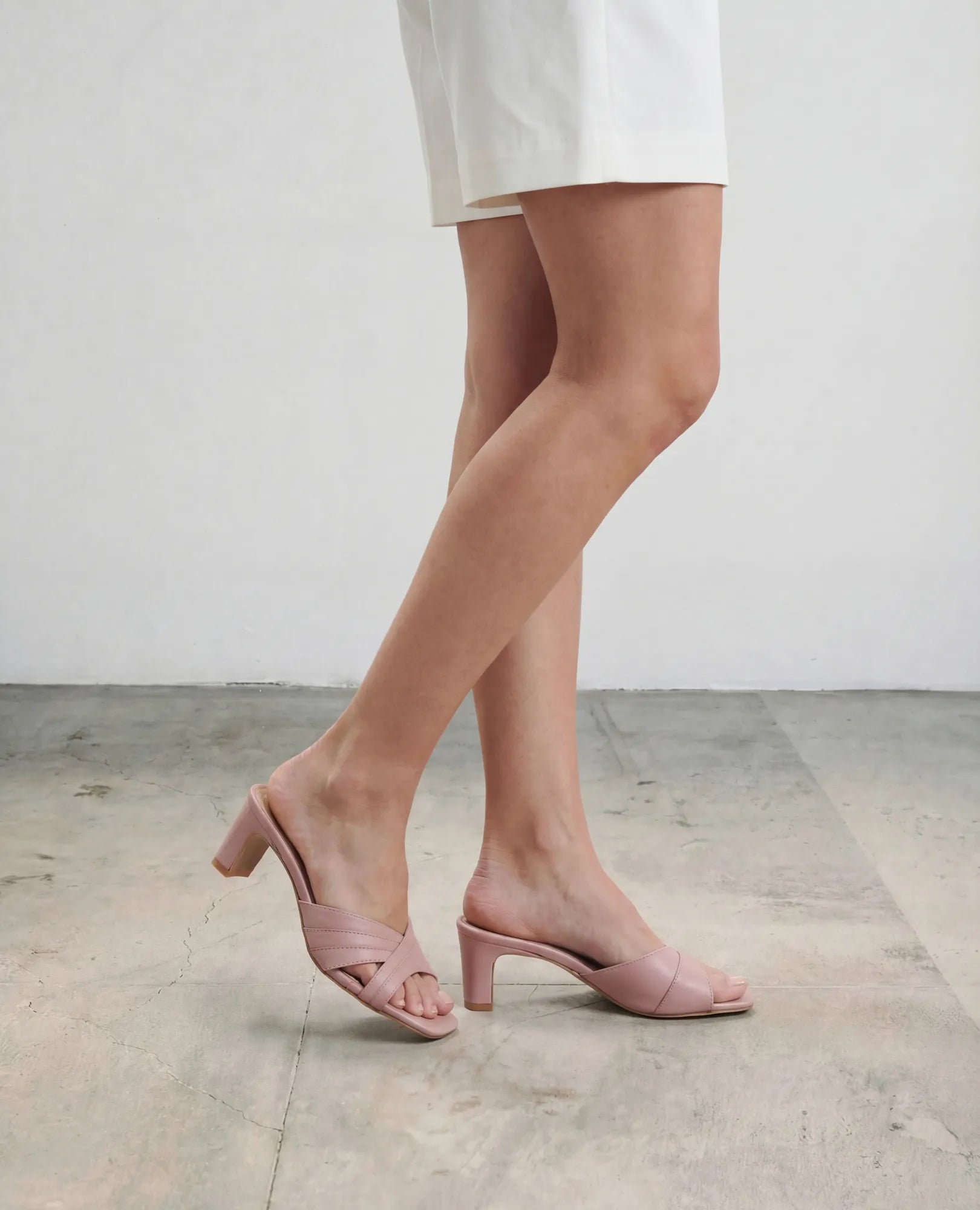 אדם לבוש במכנסיים קצרים לבנים וכפכפי עקב של B.unique בצבע ארגמן ורוד, על רצפה בהירה עם רקע אחיד.