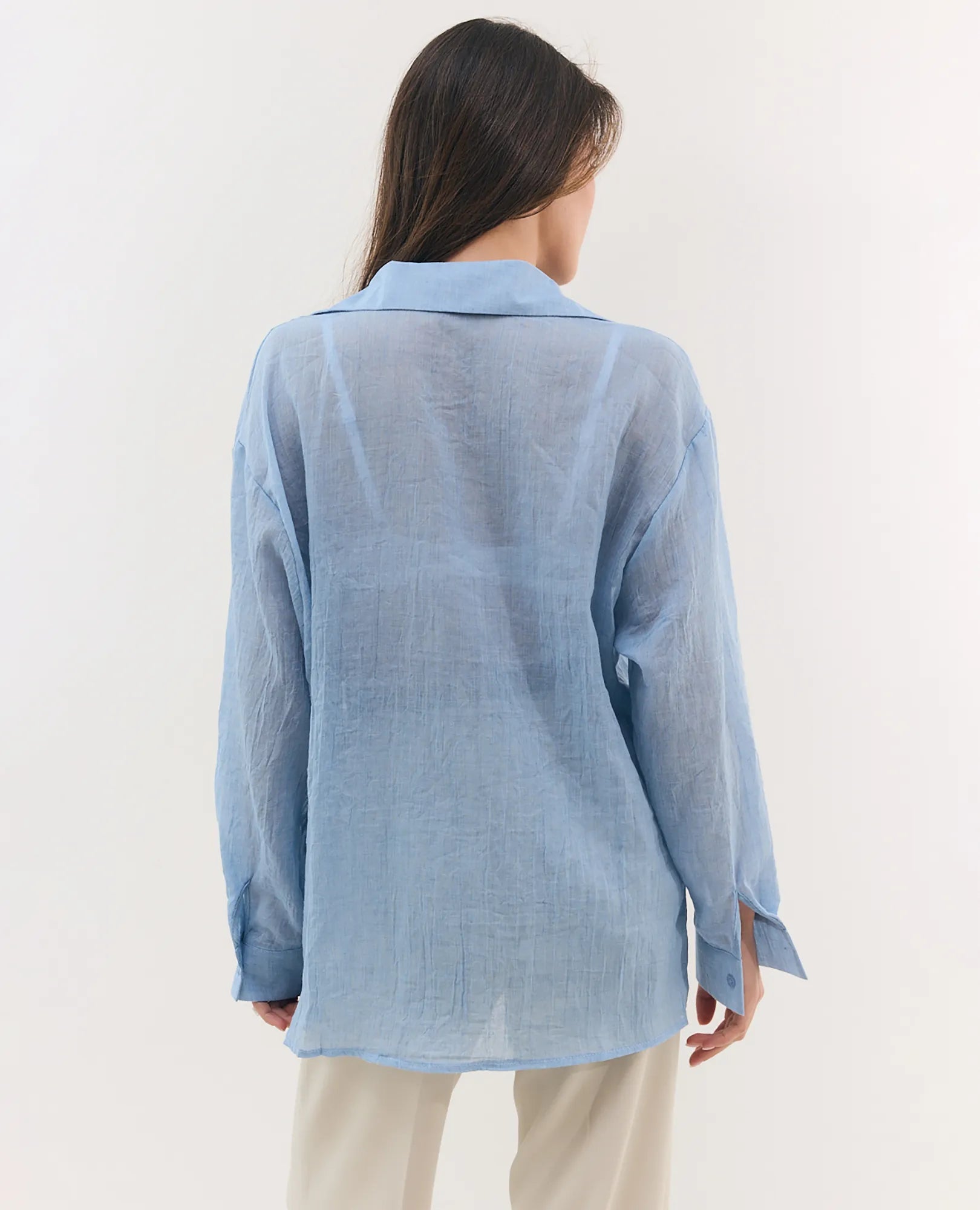 אישה עם שיער ארוך לובשת חולצה מכופתרת בצבע רובי בהיר של המותג B Unique ומכנסיים בצבע שמנת, כשגבה מופנה למצלמה.
