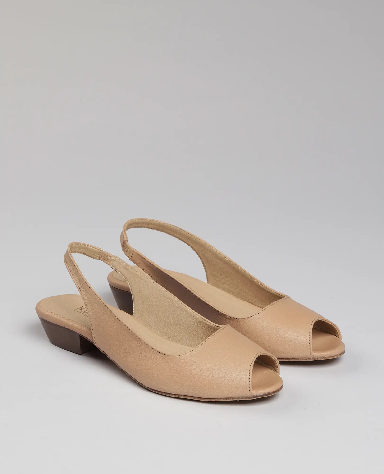 זוג סנדלי עקב-סוזי של B.unique בצבע ניוד, סנדלים עם עקב נמוך ופתח באצבעות על רקע אפור בהיר חלק.