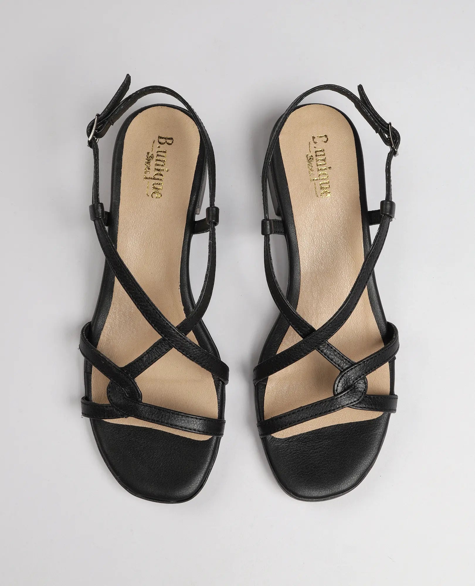 זוג סנדלי רצועות איקס-מולי מבית B.unique, מעור שחור עם רצועת קרסול ועקב נמוך על רקע לבן.