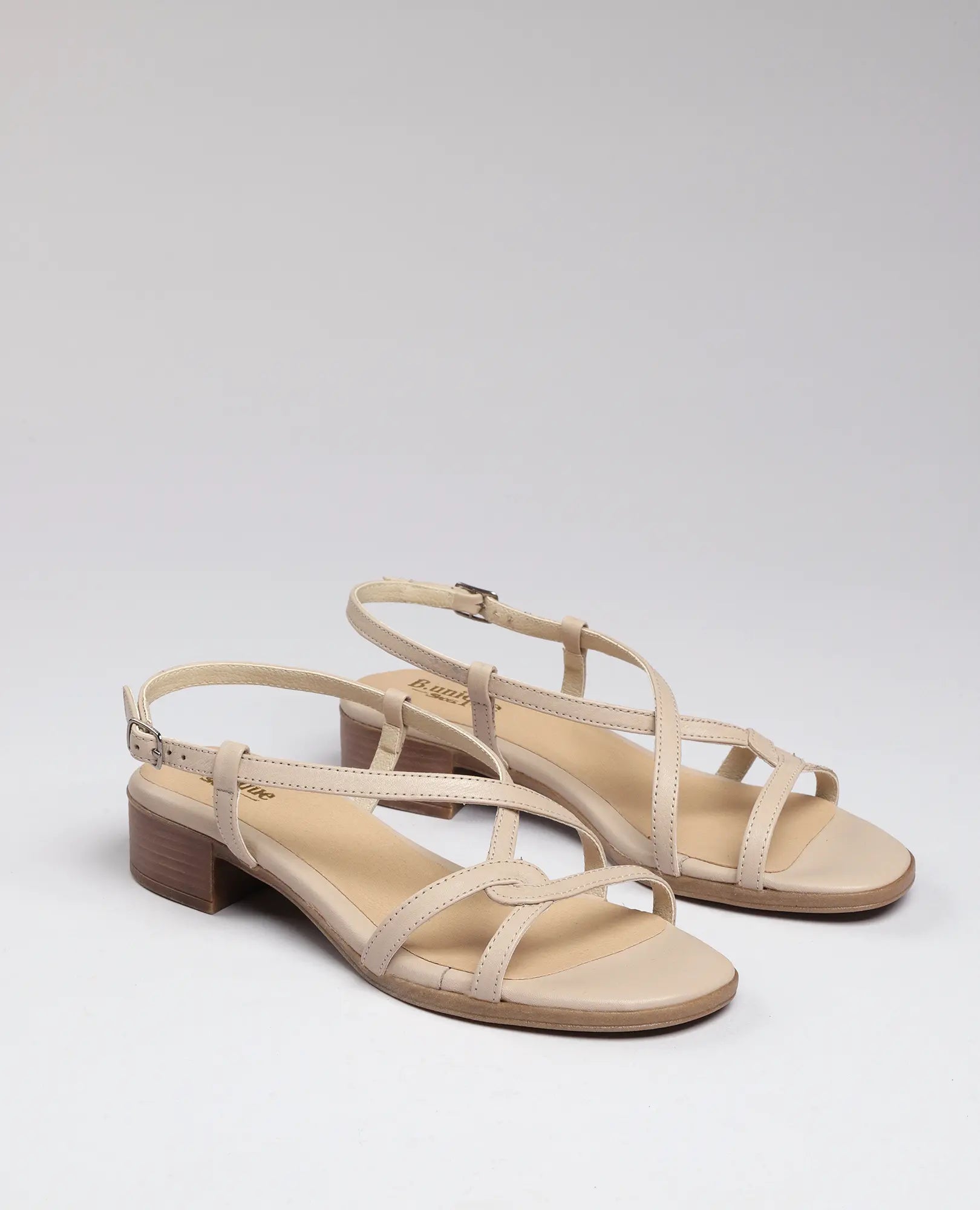 זוג סנדלי רצועות בצבע בז' מדגם "איקס-מולי" של B.unique, עם רצועות בקרסול ועקבים נמוכים מעץ על רקע חלק.