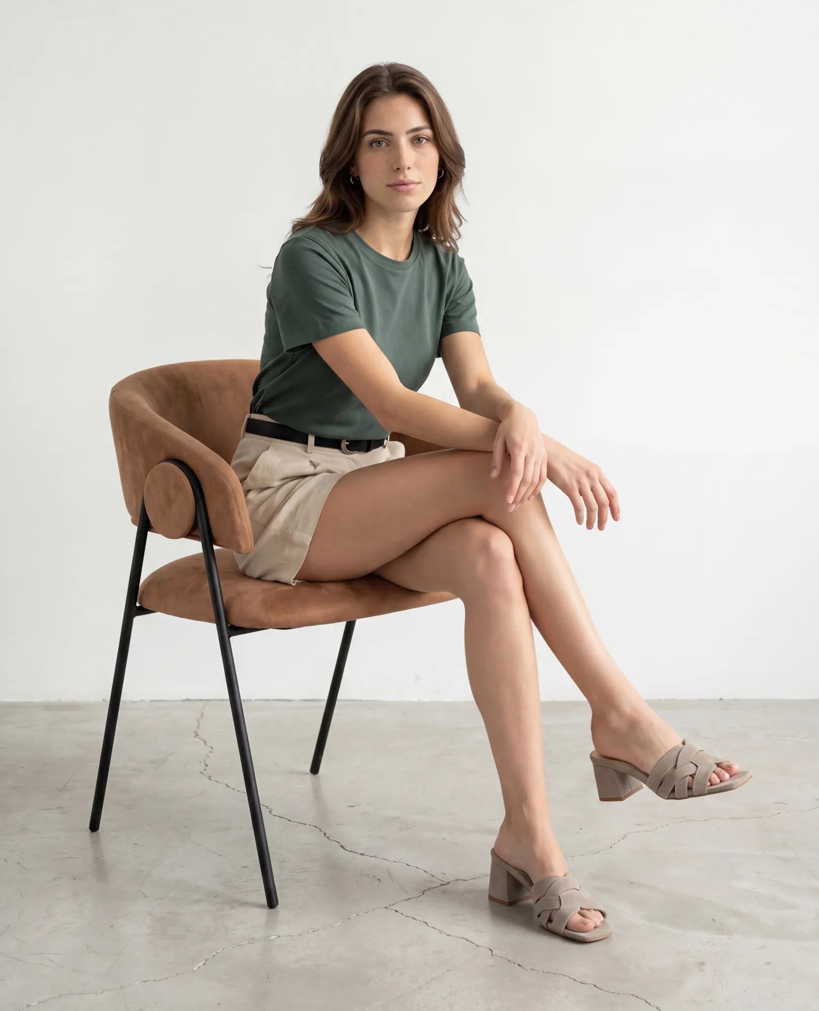 אישה לבושה בחולצה ירוקה ובמכנסיים קצרים בצבע בז' יושבת ברגליים משוכלות, נועלת כפכפי עקב-מרגי של B.unique, בחדר מינימליסטי.