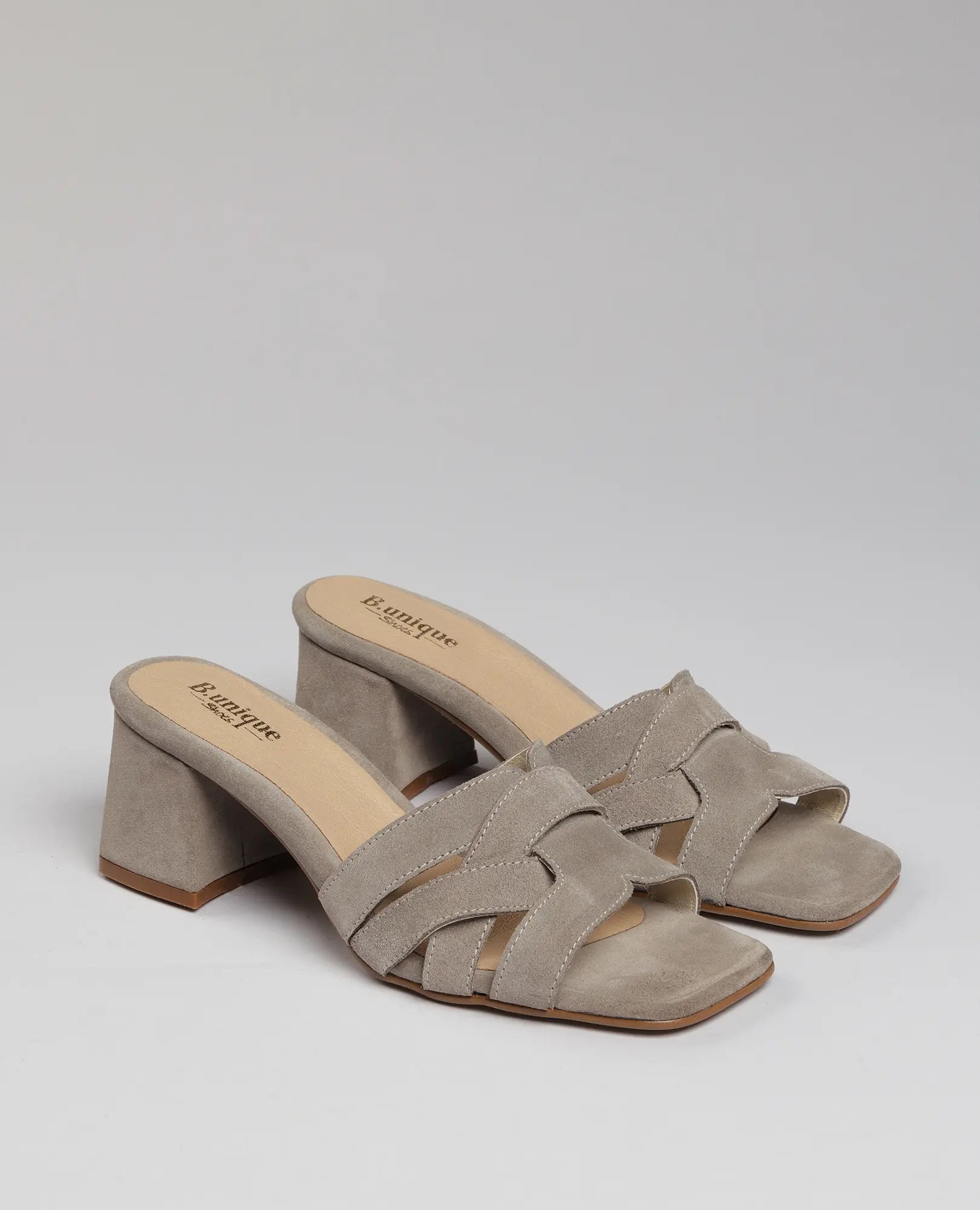 כפכפי עקב-מרגי מבית B.unique מעור זמש בצבע אבן, עם עקב רחב ורצועות קלועות, על רקע אפור חלק.