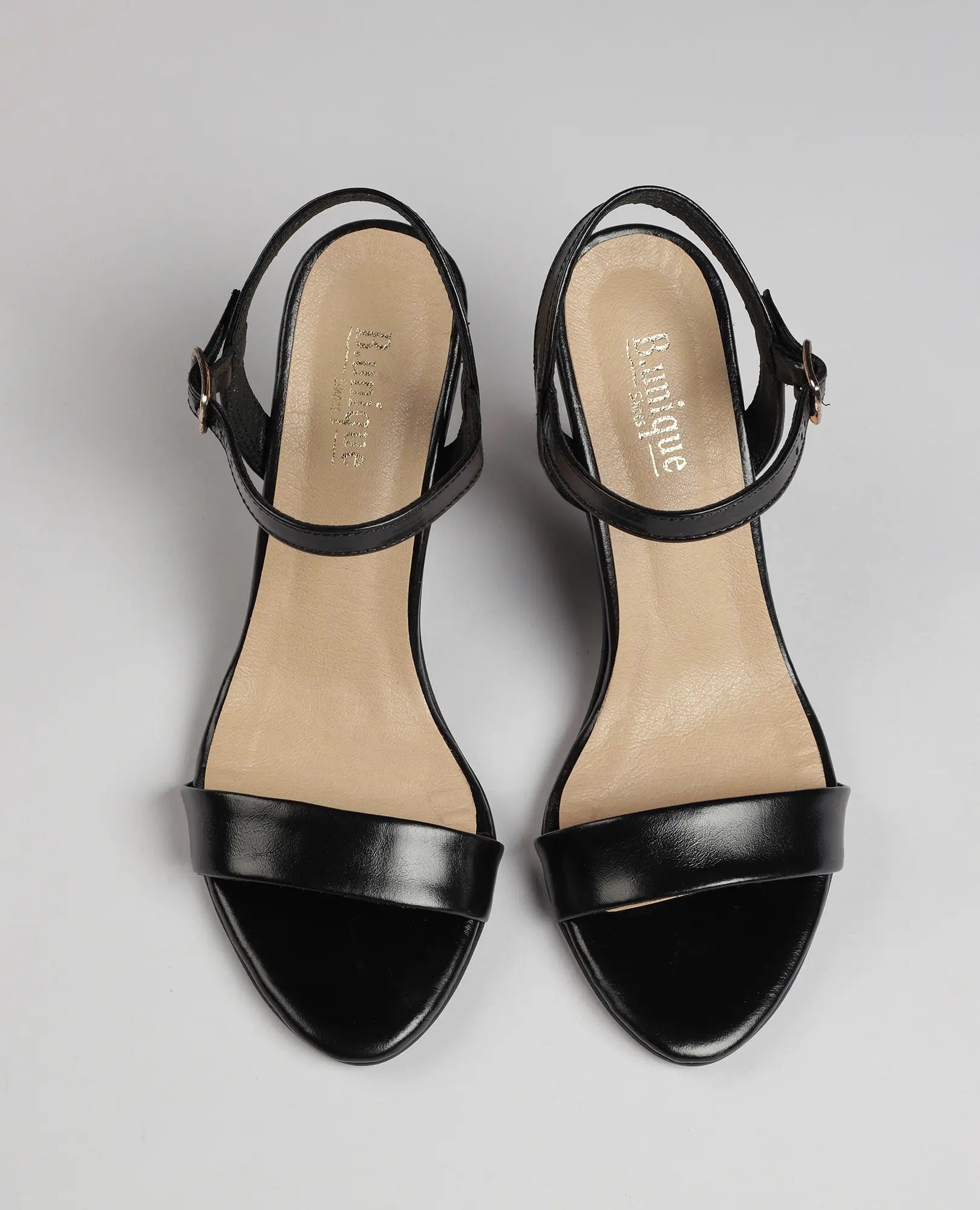 זוג סנדלי עקב גבוהים בצבע שחור מדגם "גלן" של המותג B.unique, עם רצועות בקרסול, מוצגים על רקע אפור בהיר.