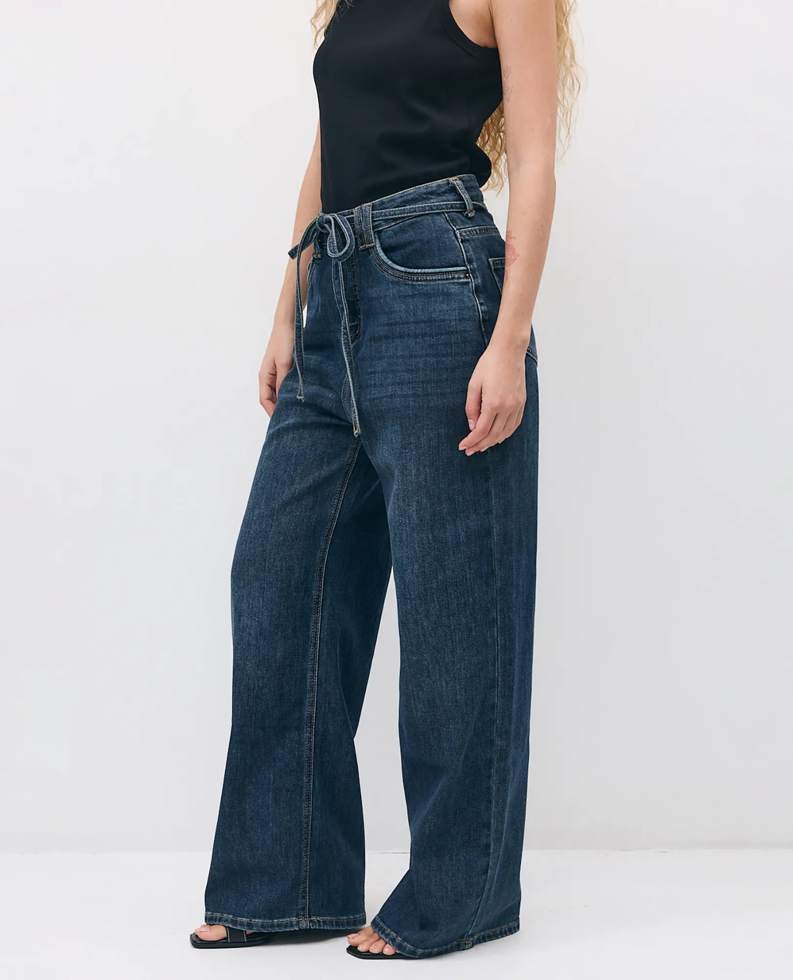 אישה לבושה בחולצה שחורה ללא שרוולים ובג'ינס נועה מבית B Unique, עומדת על רקע לבן חלק.