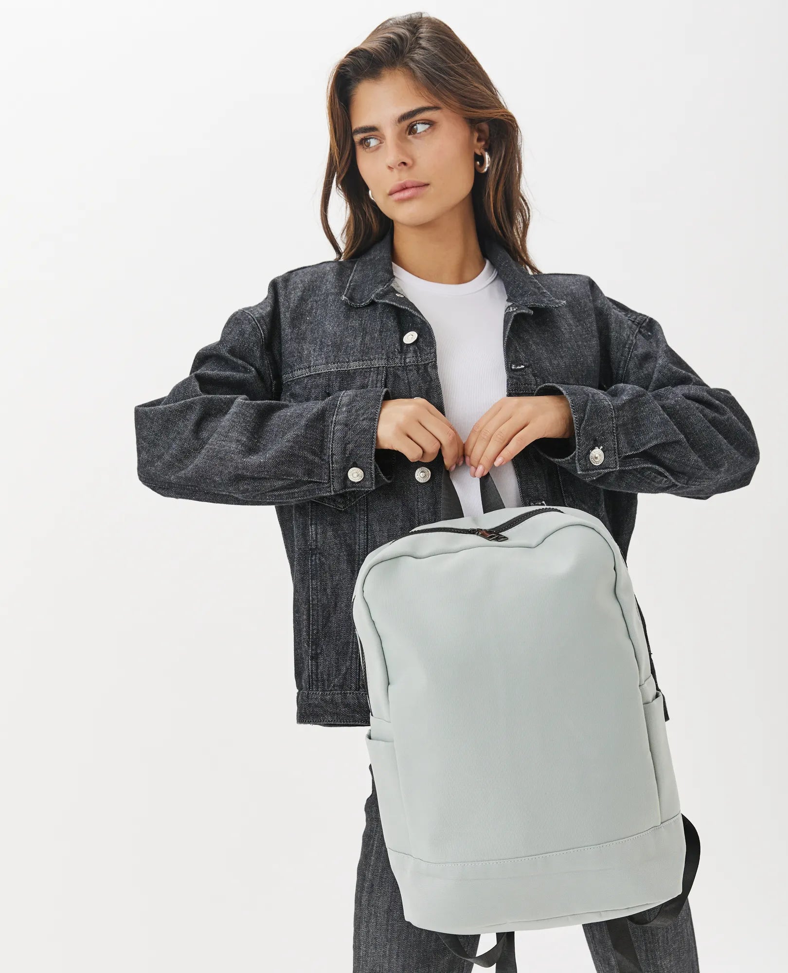 אישה עם ג'קט דנים שחור מחזיקה תיק גב- נועה של B-Unique Bags בגוון אפור בהיר על רקע לבן.