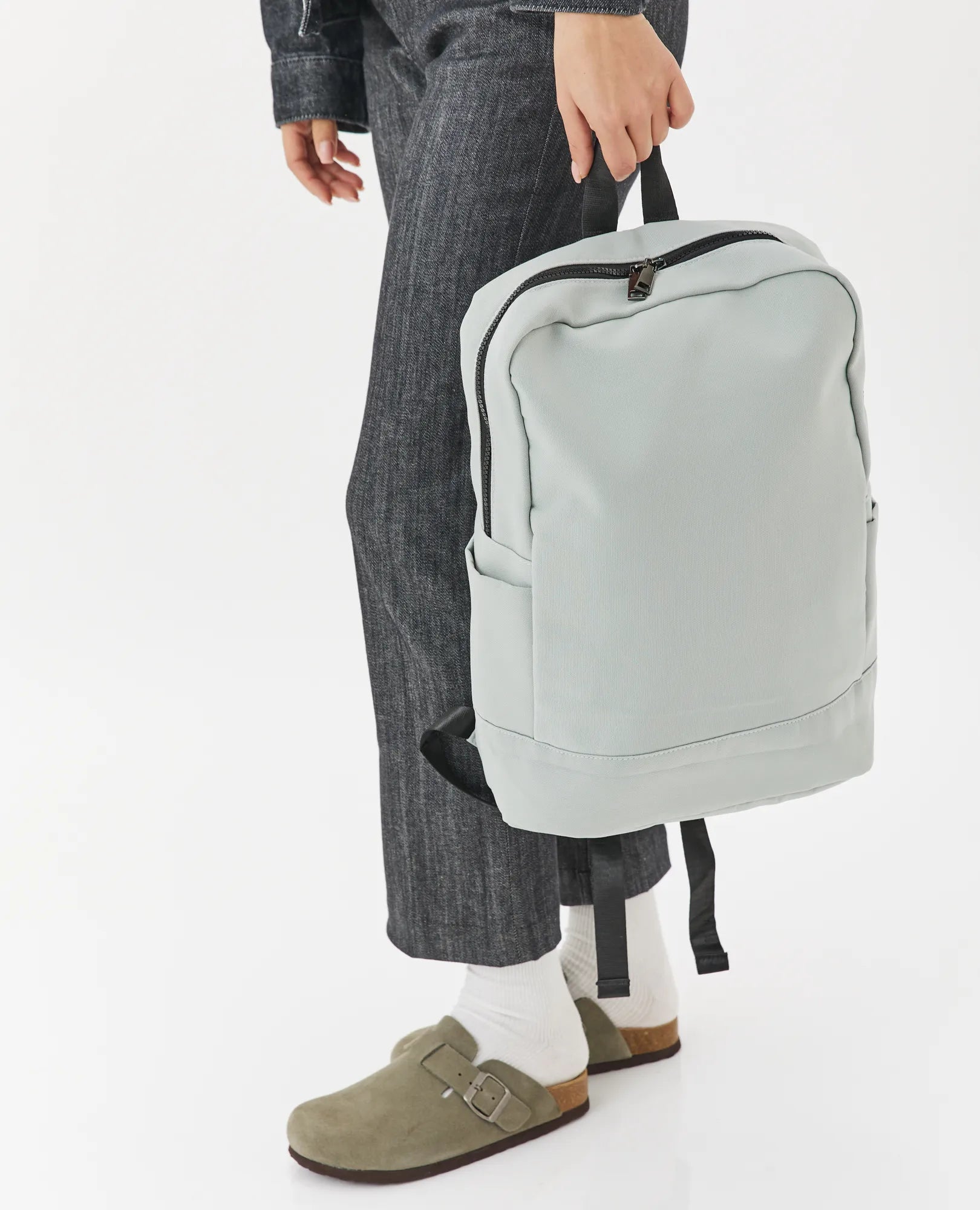 אדם לבוש במכנסיים אפורים ונעלי עץ חומות אוחז בתיק גב- נועה אפור בהיר B-Unique Bags על ידי הידית על רקע לבן.