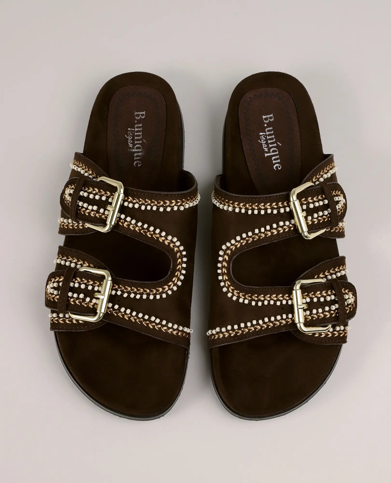 זוג כפכפים B-Unique VEGAN עם רצועות ואבזם - דגם "צור", מעור זמש חום כהה עם אבזמים מוזהבים ותפרים לבנים.