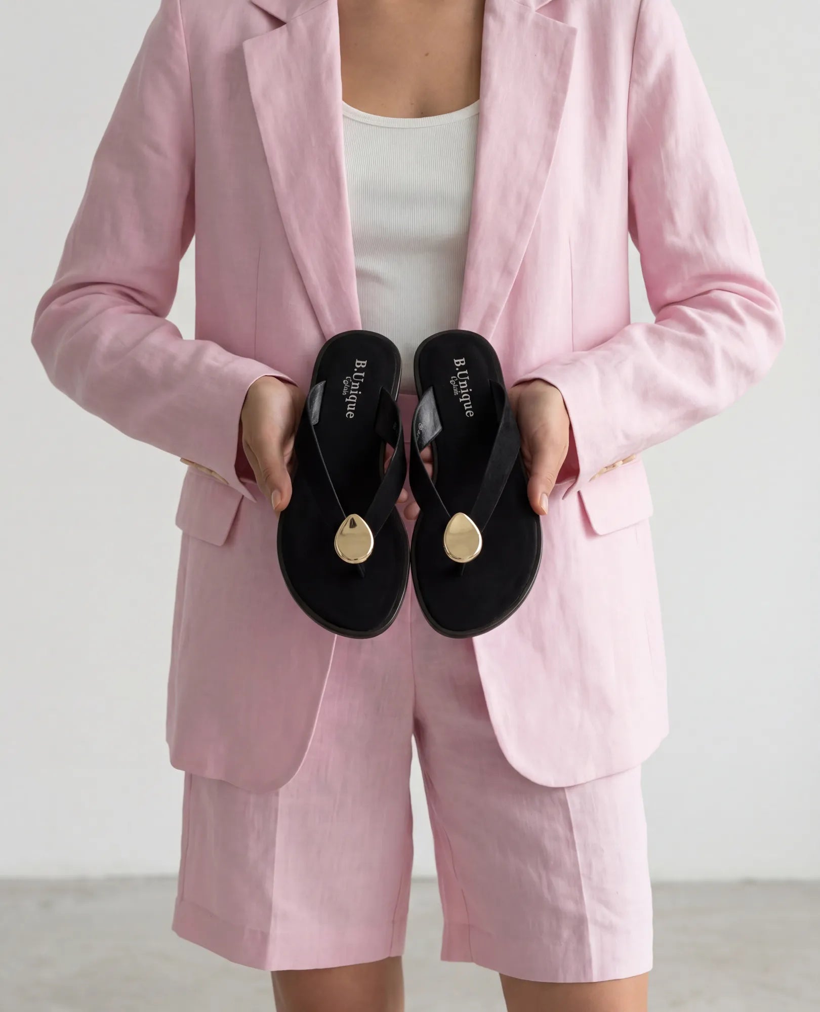 אדם לבוש בחליפה ורודה מחזיק כפכפים שטוחים מדגם "חושן" של המותג B-Unique VEGAN בצבע שחור, עם נגיעות זהב, על רקע אחיד.