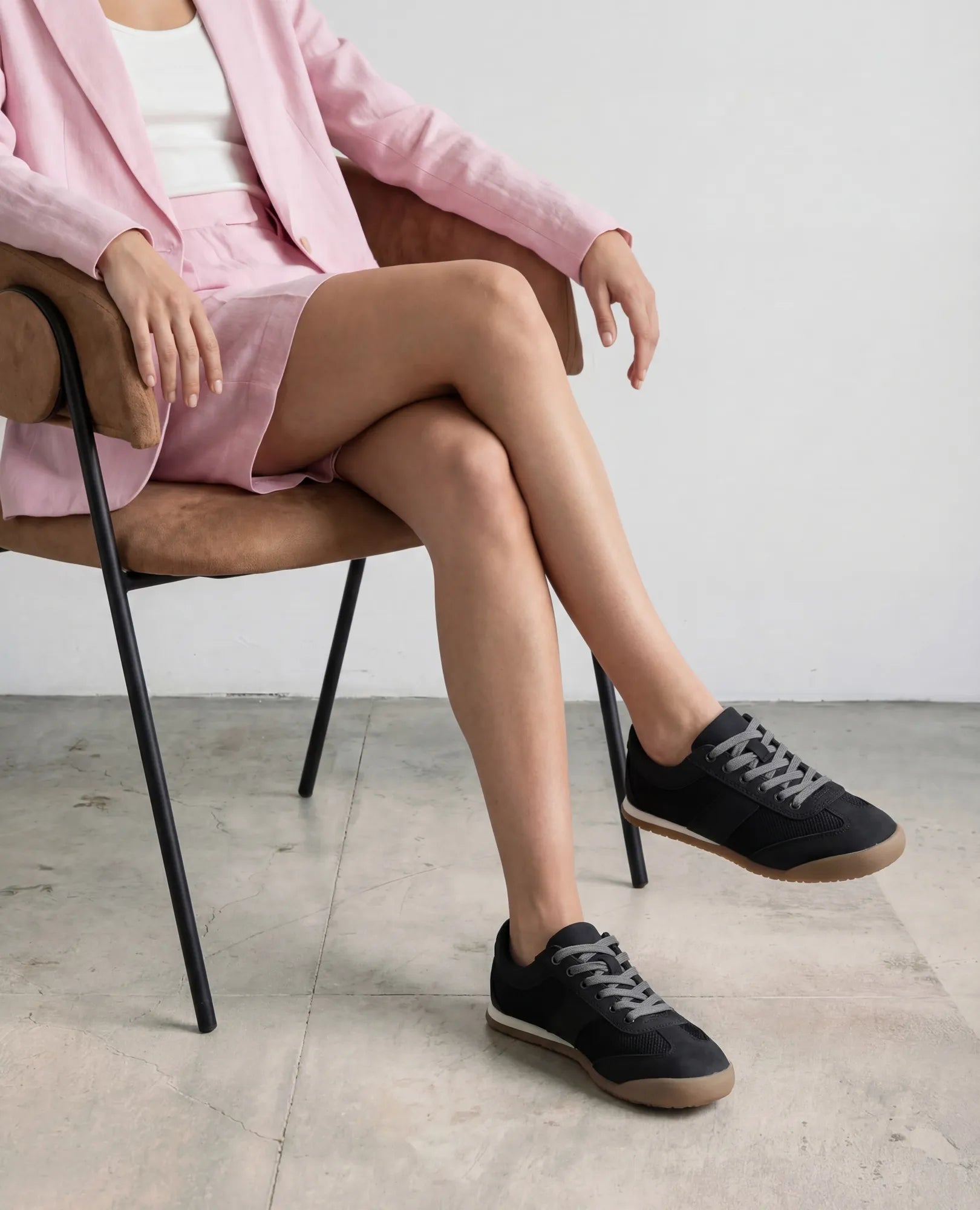 אדם לבוש בז'קט ורוד בהיר ומכנסיים קצרים יושב על כיסא, נועל נעלי סניקרס- טניה של B-Unique VEGAN על רצפת בטון.