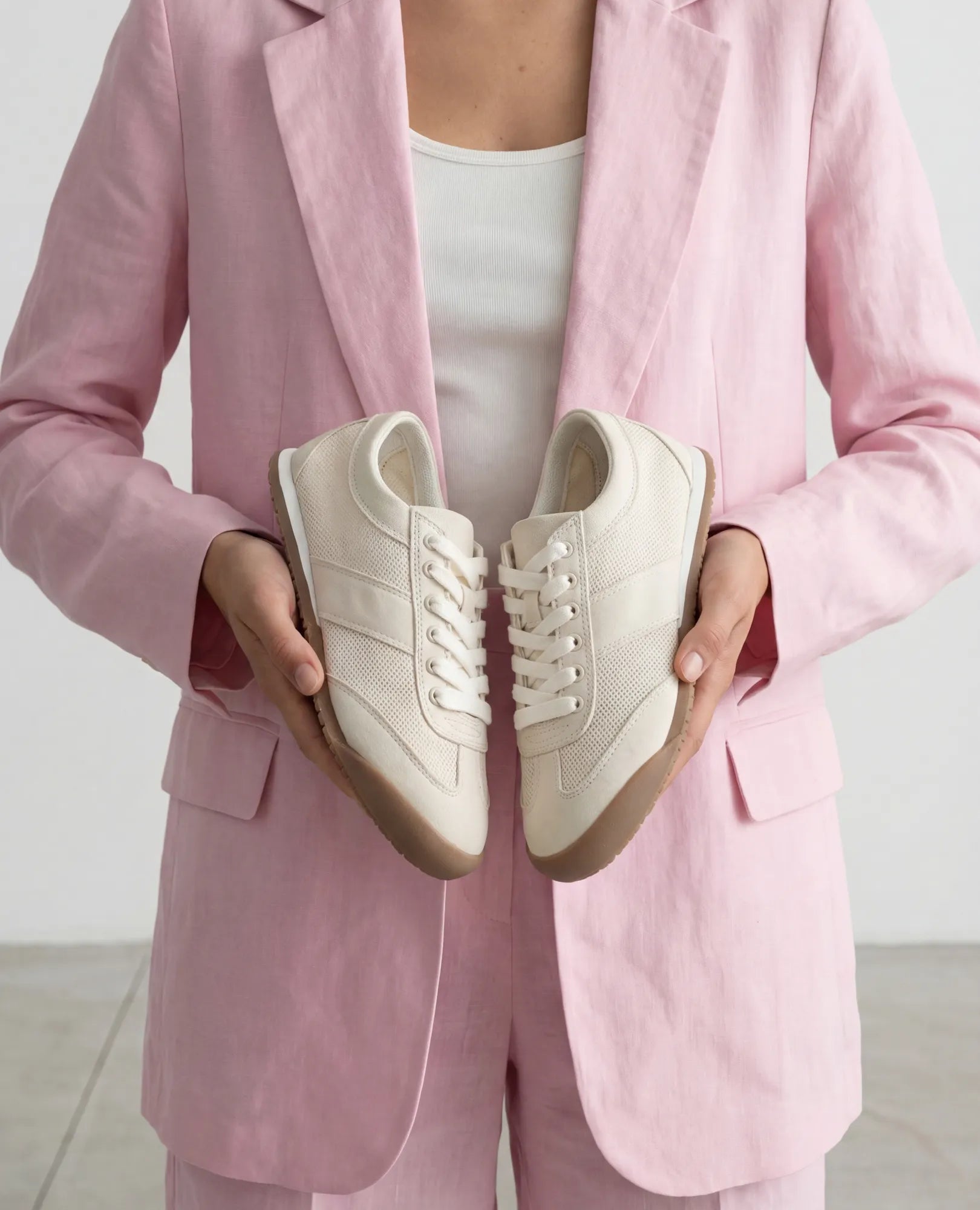 אדם בחליפה ורודה מחזיק נעלי סניקרס- טניה מבית B-Unique VEGAN, בעלות מראה זמש בז' וסוליות גומי.