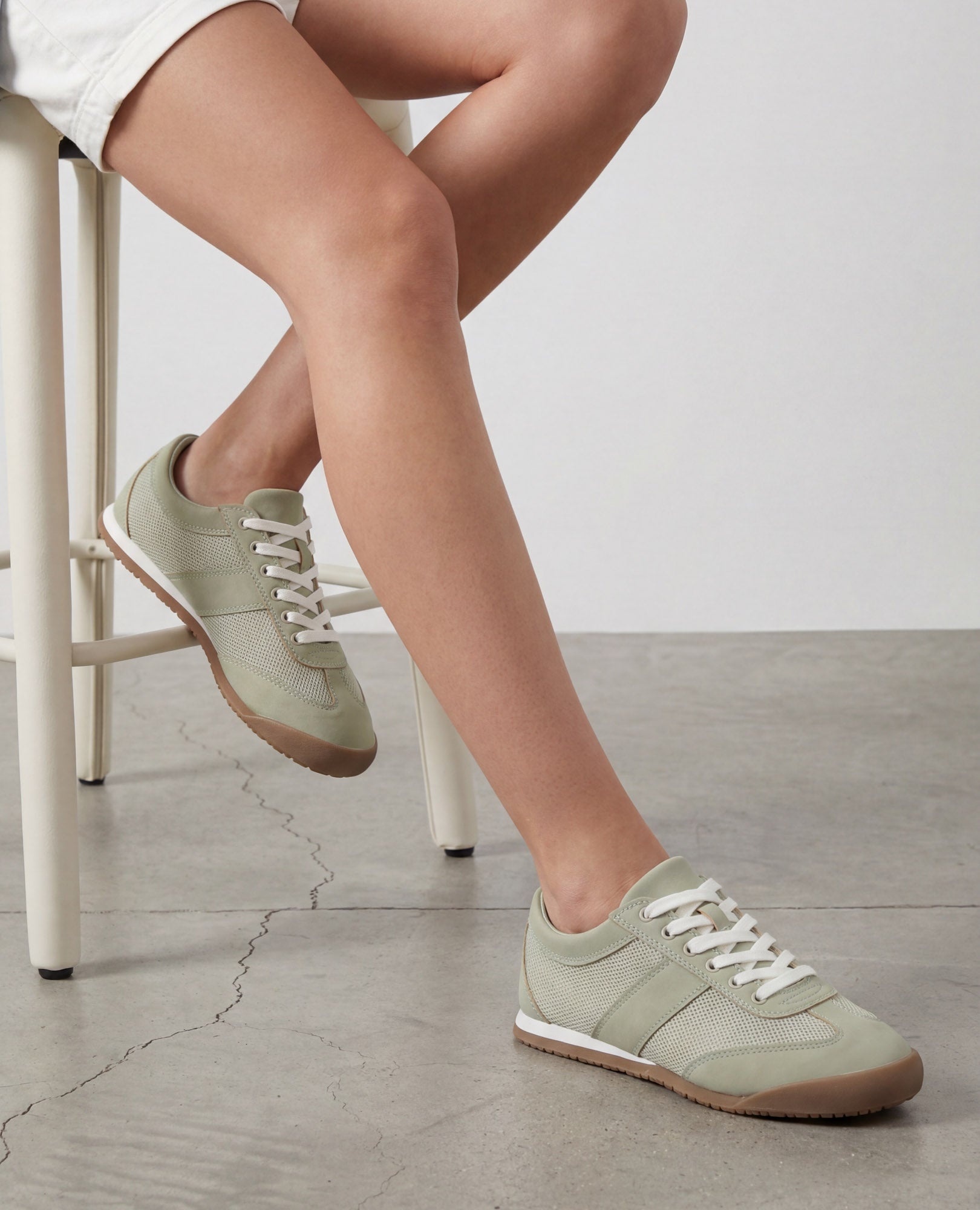 אדם יושב על שרפרף לבן, לבוש בנעלי סניקרס - טניה מבית B-Unique VEGAN בצבע ירוק זית ובמכנסיים קצרים לבנים.