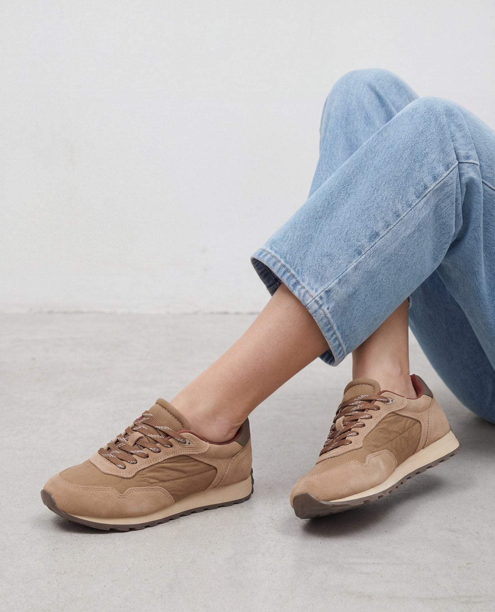 אדם לבוש בג'ינס תכלת ונעלי סניקרס עור של B.unique יושב ברגליים משוכלות על רצפה אפורה בהירה ליד קיר לבן.