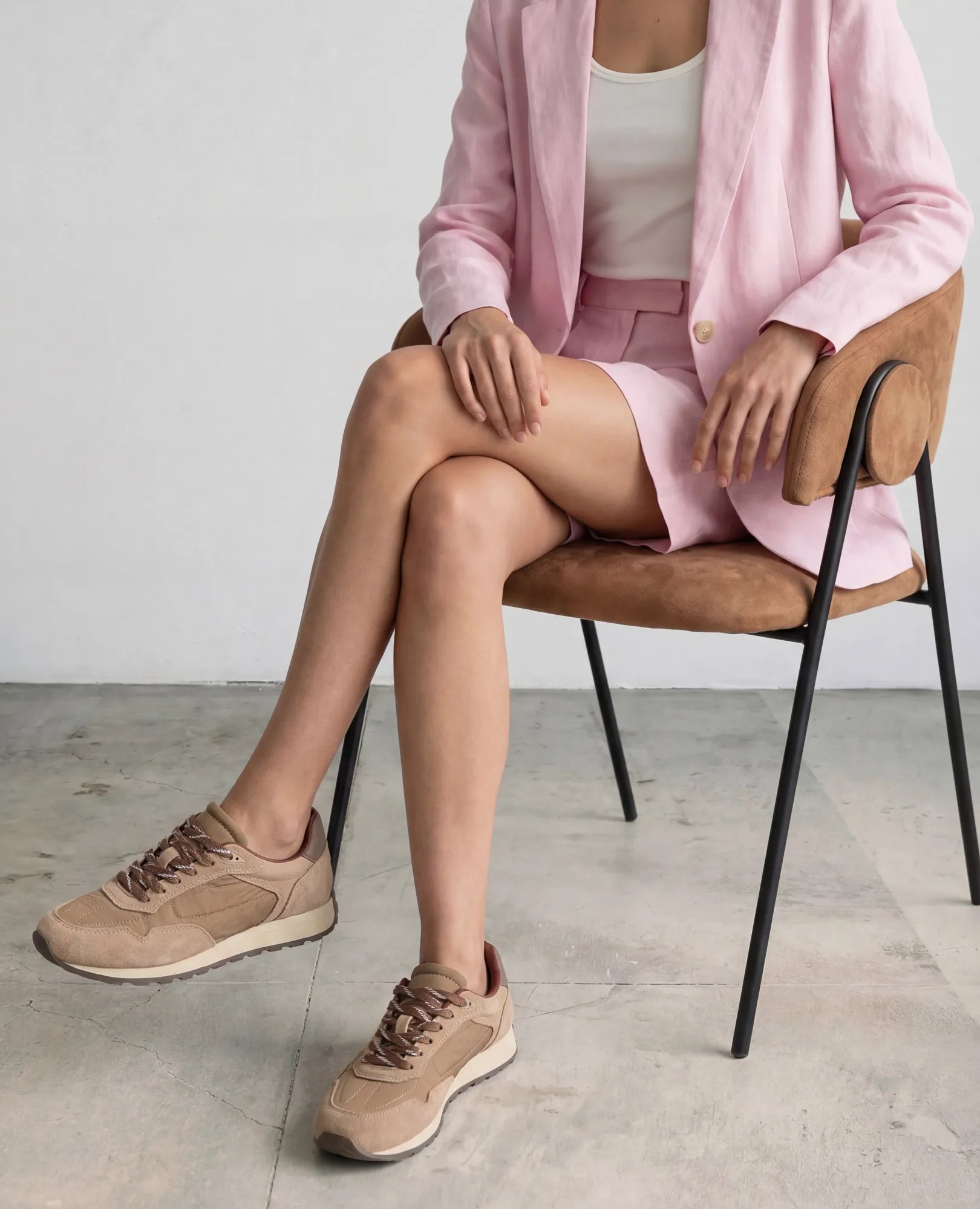 אישה לבושה בבלזר וחצאית בצבע ורוד בהיר יושבת על כיסא, נועלת נעלי סניקרס עור- ניילה של B.unique על רצפת בטון.