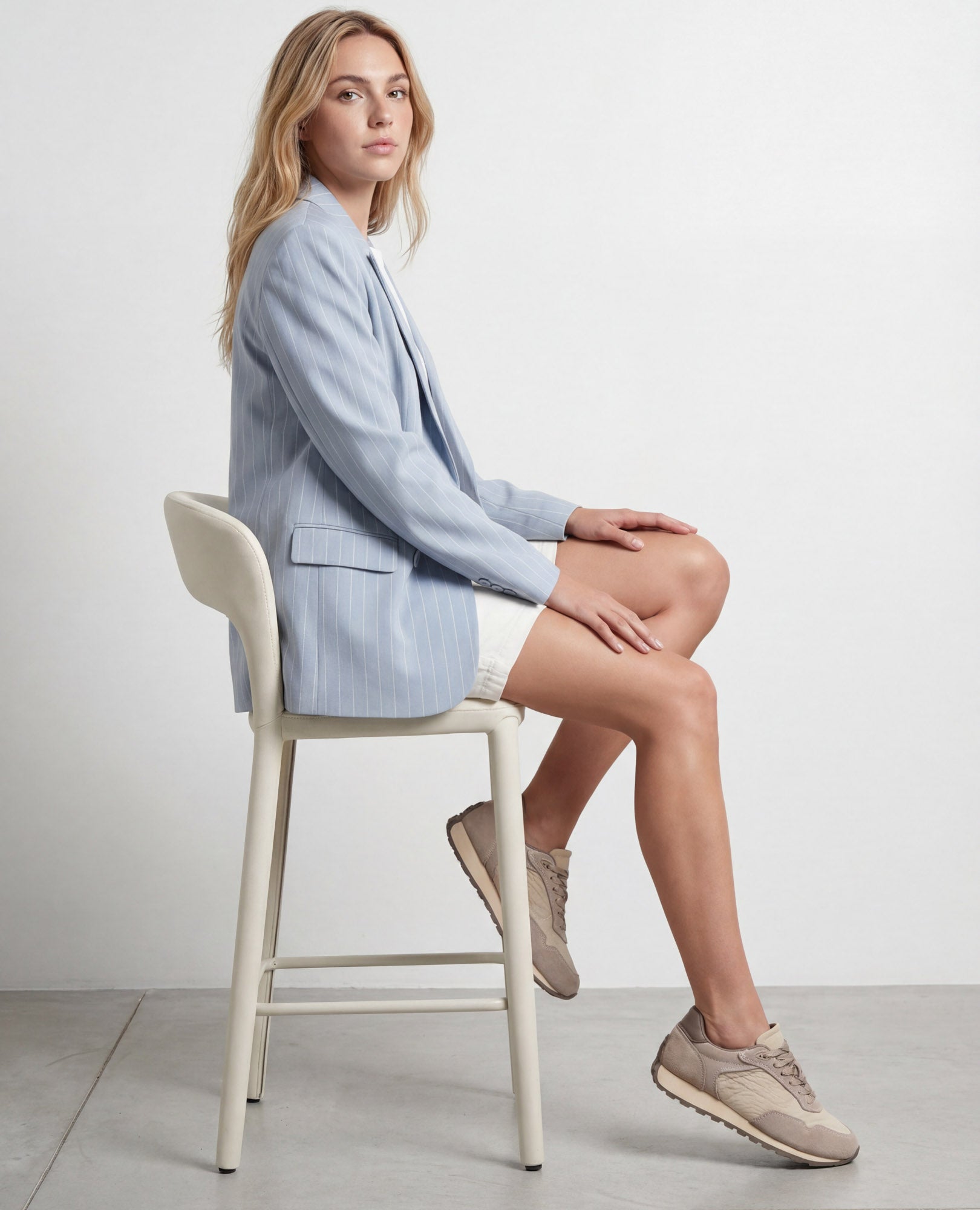 אישה בחליפת בלוזון בהירה ונעלי סניקרס עור של B.unique יושבת בצד על כיסא לבן על רקע אחיד.