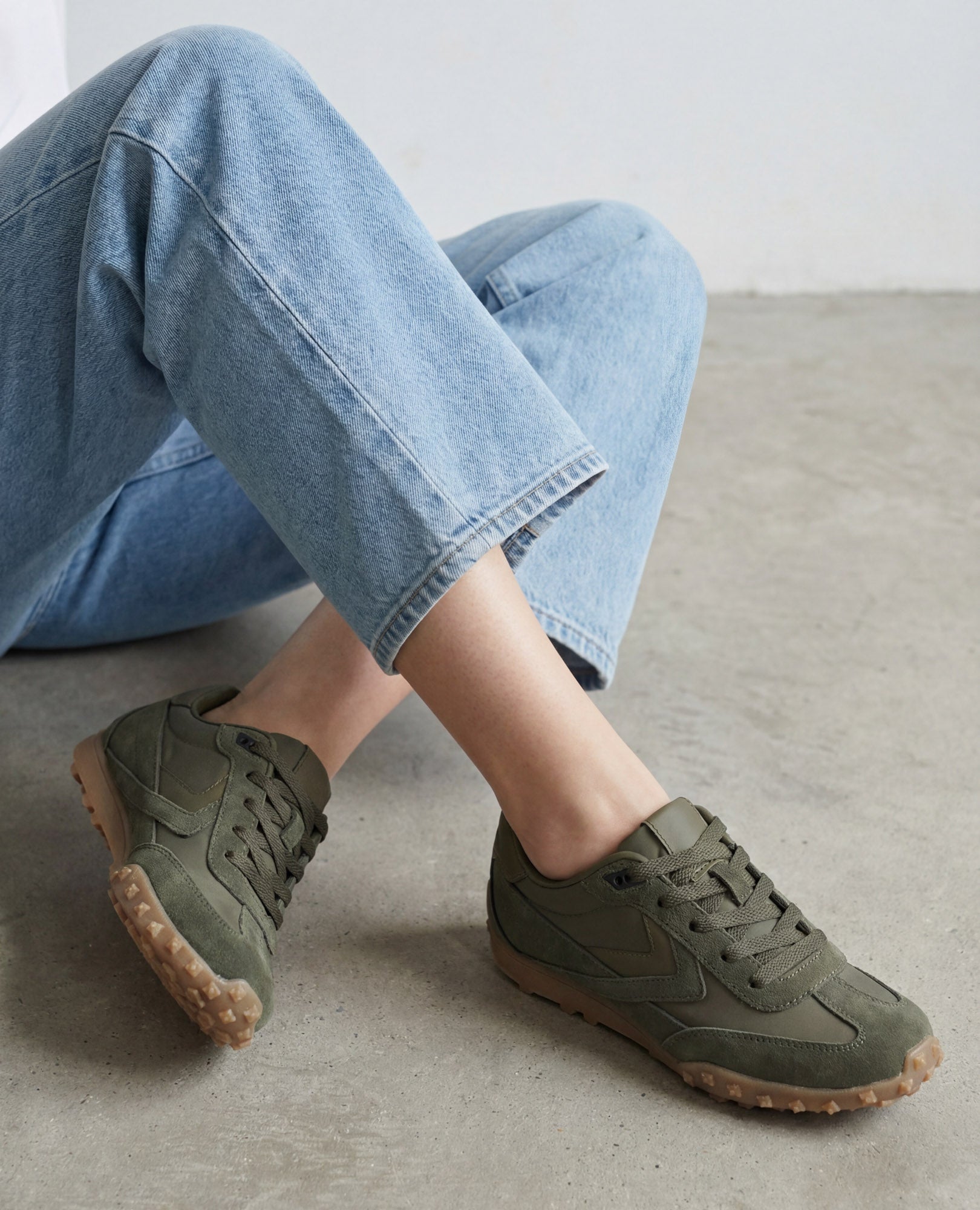 אדם לבוש בג'ינס תכלת וסניקרס מעור זמש- נטשה מבית B.unique, יושב ברגליים משוכלות על רצפת בטון.