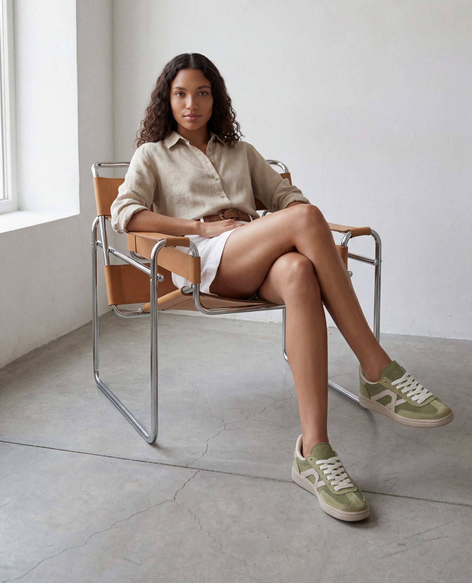 אישה יושבת על כיסא מודרני, לבושה בחולצה בצבע בז', מכנסיים קצרים לבנים ונעלי סניקרס דמוי זמש- נווה מבית B-Unique VEGAN בחדר מואר.