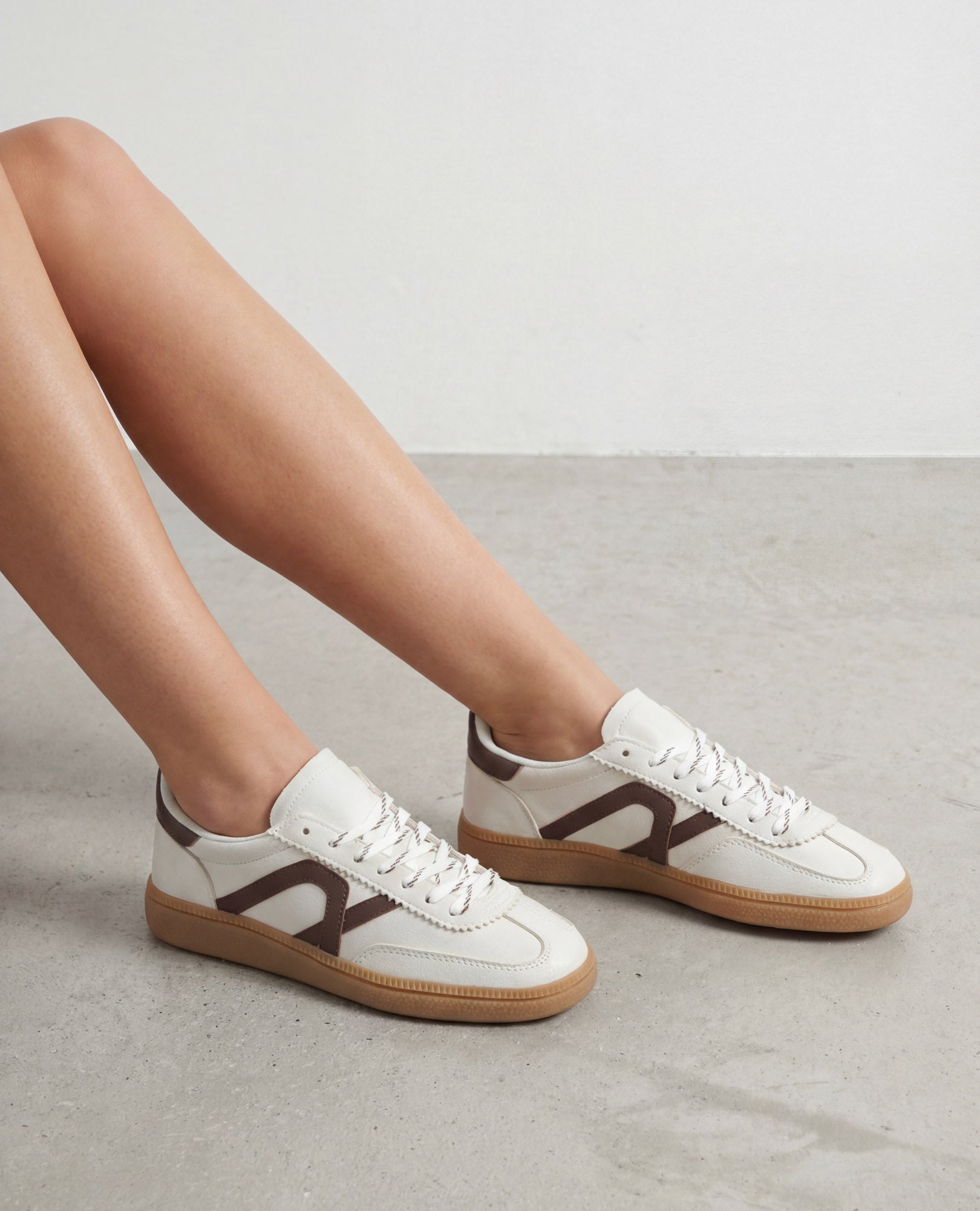 אדם נועל סניקרס דמוי זמש-נווה של B-Unique VEGAN, יושב על רצפה אפורה בהירה.