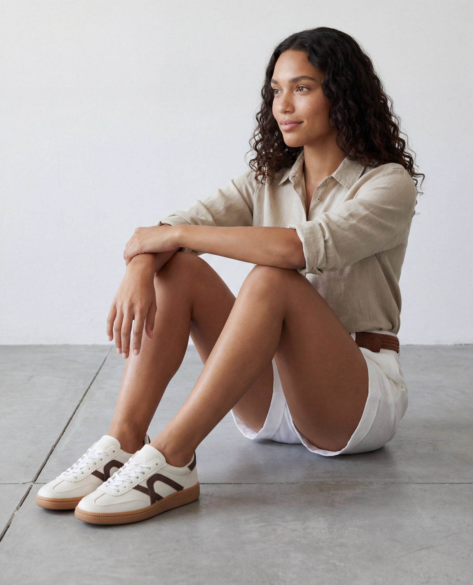 אישה עם שיער מתולתל יושבת על הרצפה, לבושה בחולצה בצבע בז' דמוי עור זמש, מכנסיים קצרים לבנים וסניקרס דמוי זמש- נווה של B-Unique VEGAN.