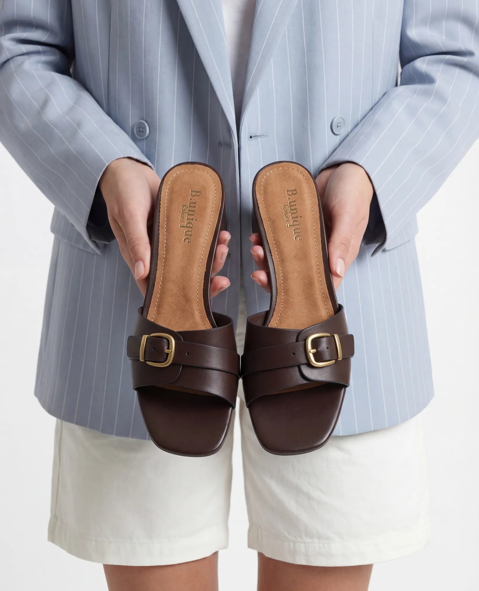 אדם לבוש בבלזר תכלת ומכנסיים קצרים לבנים, מחזיק מול המצלמה כפכפי עקב-שחק של המותג B-Unique VEGAN בצבע קפה.