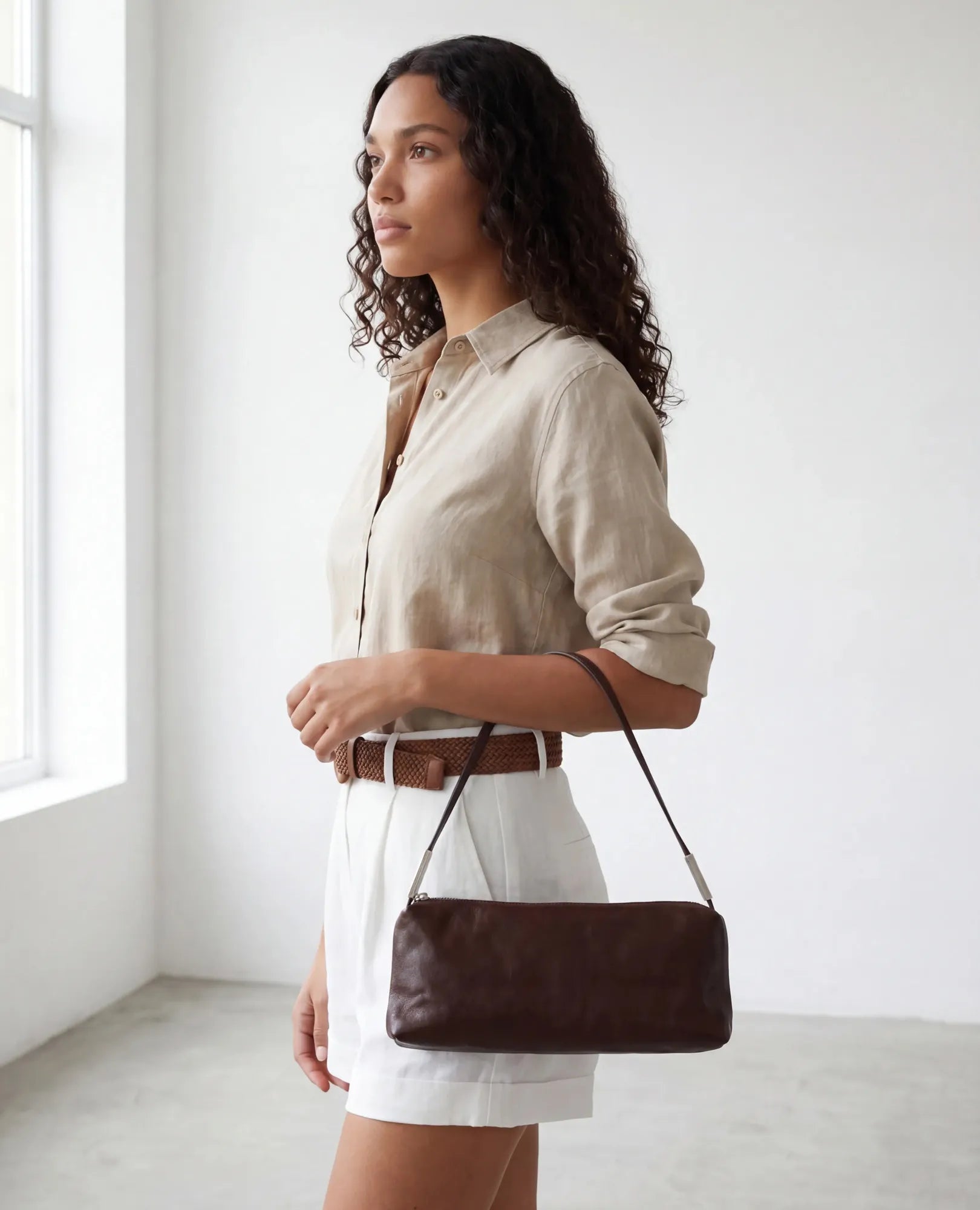 אישה עם תיק צד מלבני מדגם "אליס" של B-Unique Bags מונח על הכתף, לובשת חולצה בצבע בז' ומכנסיים לבנים.