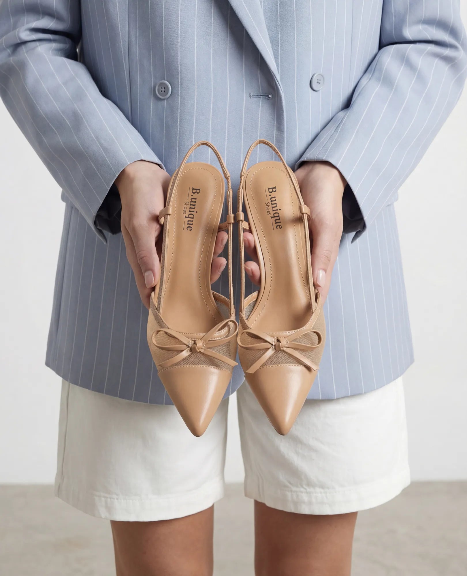 אישה לבושה בבלזר כחול עם פסים דקים ומכנסיים קצרים לבנים, אוחזת בנעלי עקב עם קצה מחודד בצבע ניוד של המותג B-Unique VEGAN.