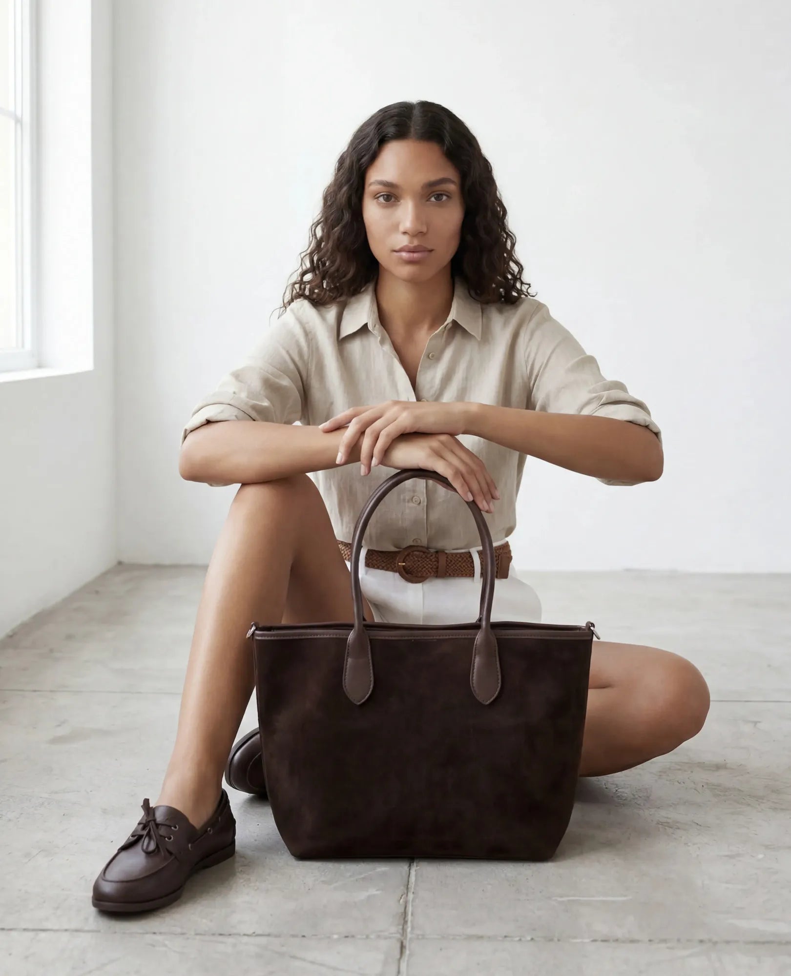 אישה עם שיער מתולתל יושבת על הרצפה, ידיה מונחות על תיק צד מזמש מעור - קמיל מבית B-Unique Bags, בחדר מואר בסגנון מינימליסטי.